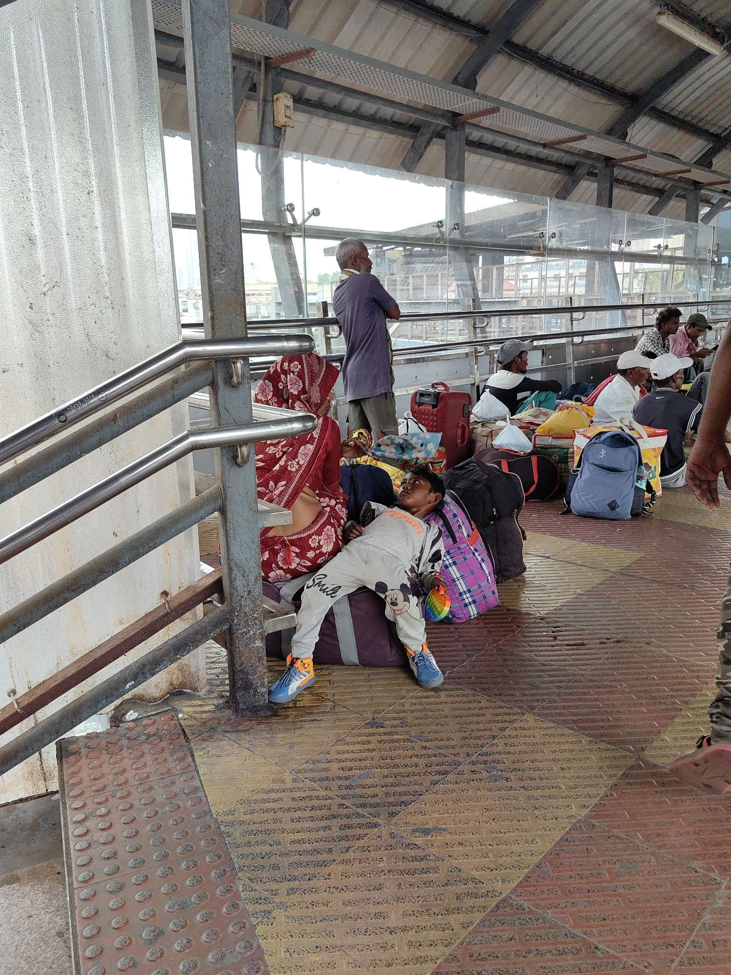 The image depicts a scene at a Mumbai, India train station, likely a platform or waiting area. In the foreground, a young boy is lying down, propped up against a large duffel bag, with his head resting on a patterned bag. He is wearing light-colored pants with Mickey Mouse graphics and blue and orange sneakers. Beside him are other bags, including a red suitcase and a blue backpack.
In the background, several people are seated on the ground or on their luggage, appearing to be waiting. A woman in a red and white floral sari is seated near a metal railing. Further back, a man in a purple shirt is standing. The environment appears to be an outdoor or semi-outdoor covered area, with a corrugated metal roof supported by metal beams. The floor is tiled in a diamond pattern. The overall impression is one of people traveling or waiting for transport in a busy urban setting.