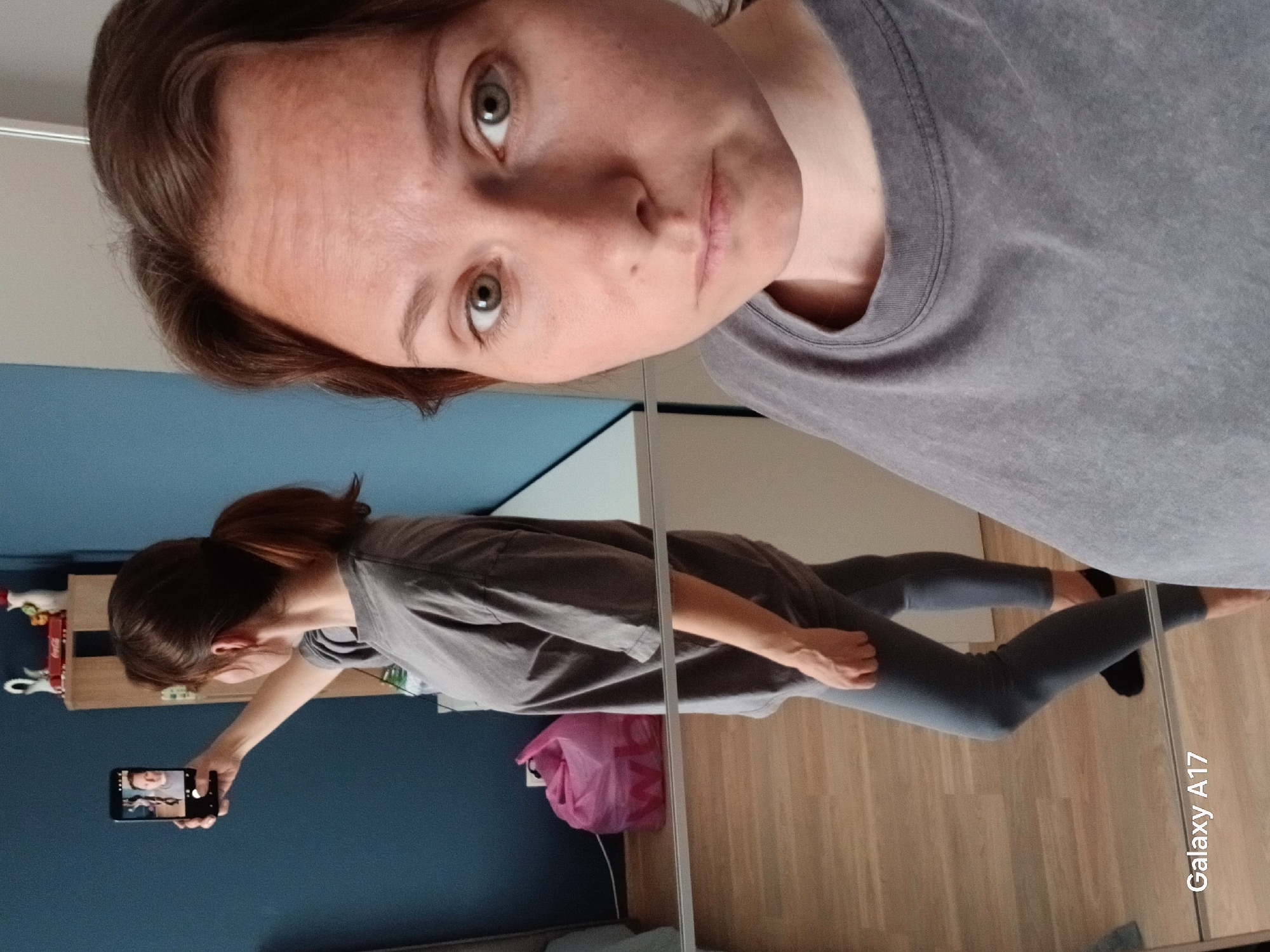 A woman is taking a selfie in front of a mirror. The mirror reflects the woman holding a phone in her left hand, angled slightly downward. She is wearing a gray t-shirt and gray leggings. The background includes a blue wall, a pink bag, and a wooden floor. The location is likely indoors, but the city of Yekaterinburg, Russia, cannot be determined from the image. A phone brand is listed in the bottom right of the image "Galaxy A17".