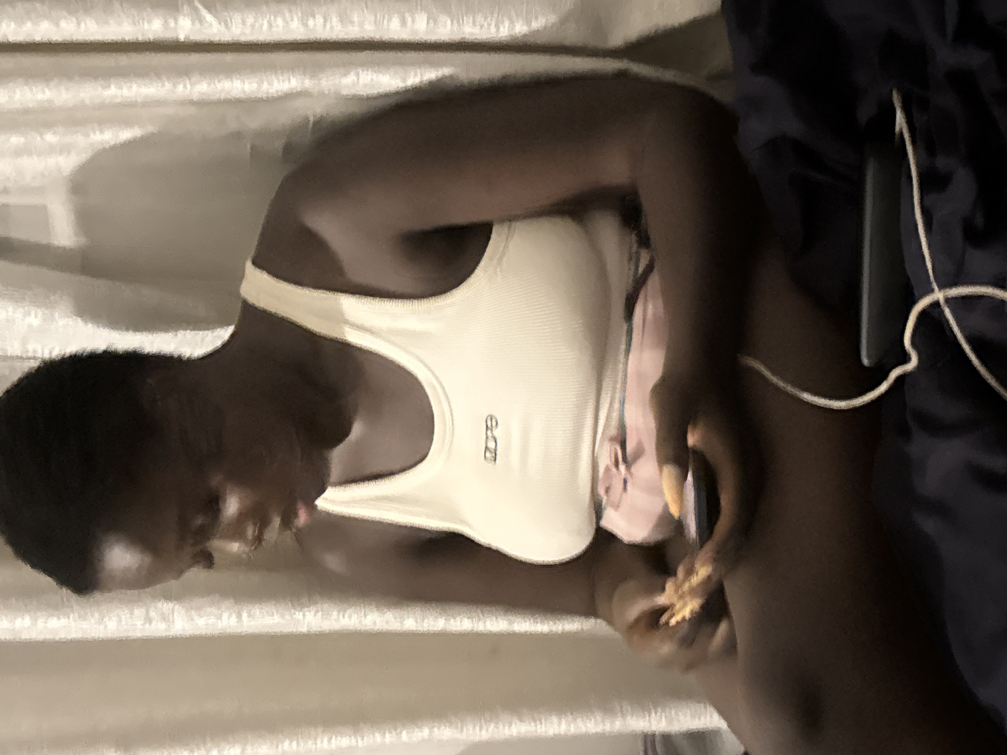 A dark-skinned woman with short hair sits on a dark blue fabric, possibly a bed or sofa, in front of a beige curtain. She is wearing a cream-colored ribbed tank top with the word "dope" embroidered on the chest in black thread. Her hands are engaged with a small object, possibly food, and a black mobile phone. The phone is partially obscured by her hands and rests on her lap. A thin white cord, possibly from earphones, runs from the phone and trails across the blue fabric. The woman's head is tilted downwards, and her expression is focused. The environment appears to be indoors, with soft lighting creating shadows on the curtain behind her.