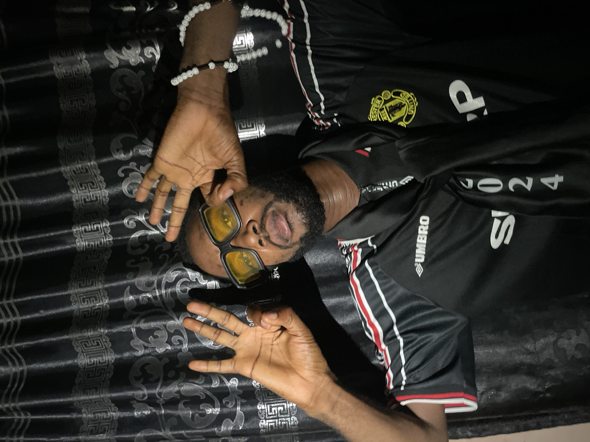 The image depicts a medium shot of a Black man standing in front of a dark, patterned curtain, likely in Lokoja, Nigeria. The man is wearing a black Manchester United jersey with red and white stripes on the sleeves and collar. He has on yellow-tinted glasses and is holding his hands up, with his fingers in a gesture. His right hand is extended, displaying four fingers, while his left hand is positioned near his face, obscuring part of his head. He has a beard and is looking towards the camera with his mouth slightly open. On his left wrist, he wears multiple beaded bracelets, predominantly white. The background consists of a black curtain with a subtle silver-toned repeating pattern of geometric shapes and floral motifs. The lighting in the image is dramatic, casting shadows on the man's face and the curtain.