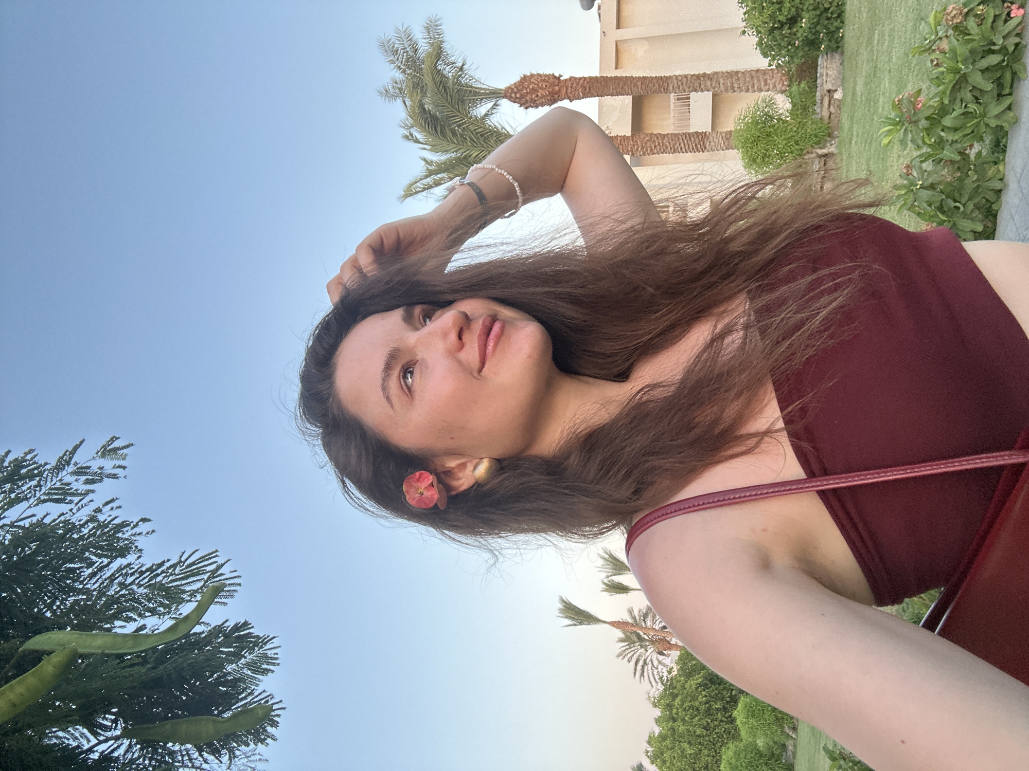 In Hurghada, Egypt, a young woman with long, dark brown hair, adorned with a small red flower behind her right ear, poses for a selfie outdoors. She is wearing a maroon strapless top and a light-colored bracelet on her left wrist, with a purse strap visible across her chest. Her hand rests on top of her head as she looks to the side. The background features lush green vegetation, palm trees, and a light-colored building under a clear blue sky, suggesting a resort or hotel setting.