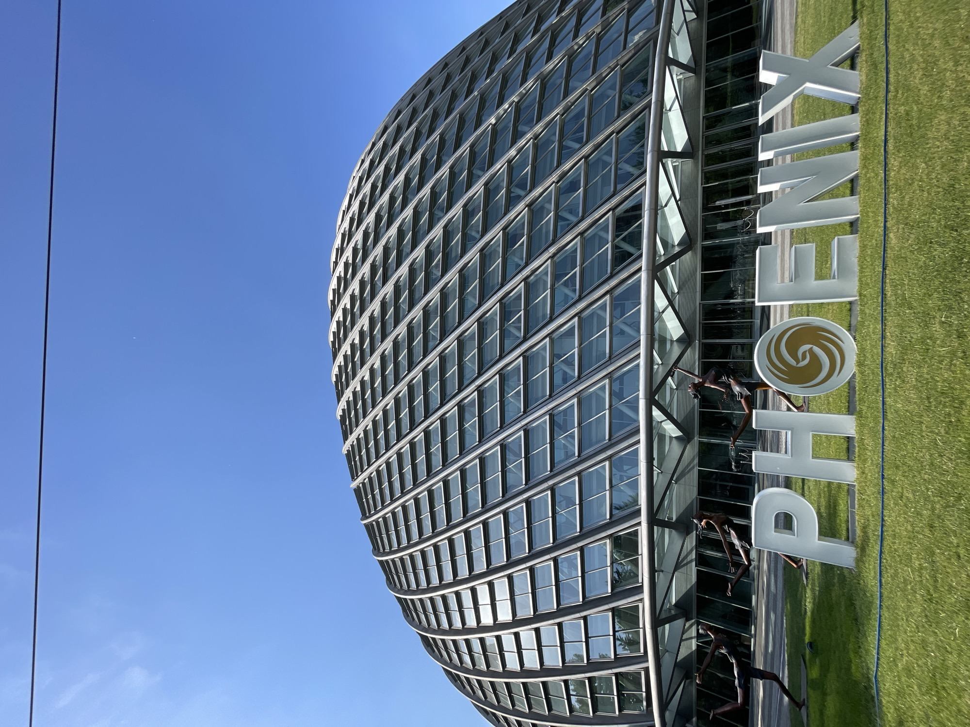 The image shows the Phoenix International Media Center in the Chaoyang District of Beijing, China. The building is a curved, metal-clad structure with numerous rectangular windows. In front of the building is a large sign that spells out "PHOENIX", complete with the company's logo. Three statues of people performing various activities are positioned adjacent to the sign and the base of the building. The surrounding area consists of green grass and a clear blue sky.