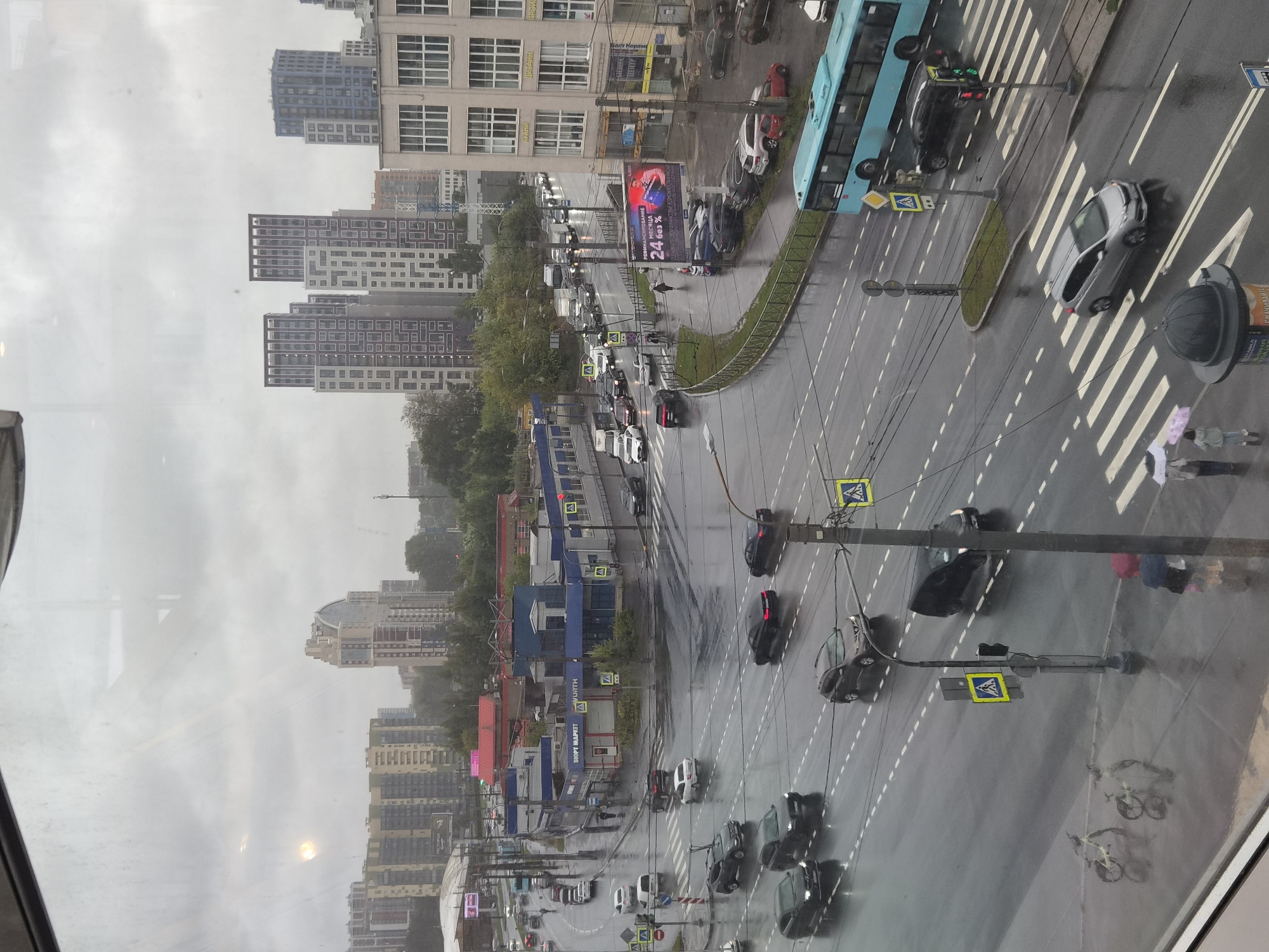 This image captures a rainy day scene in a city, likely Saint Petersburg, Russia, characterized by its overcast sky and wet roads. The perspective is from an elevated viewpoint looking down on a busy intersection with multiple lanes of traffic.
In the foreground, a light-colored minivan crosses a pedestrian crossing. To the left, two bicycles are parked on the sidewalk. Further back, a bright blue bus is moving through the intersection, with a black SUV driving behind it and crossing the pedestrian walkway. Traffic lights are visible, with one displaying red and green signals.
The mid-ground shows a complex intersection with cars navigating wet asphalt. Various vehicles, including sedans, SUVs, and a truck, are present. Pedestrians with umbrellas are visible walking on sidewalks. Buildings line the sides of the road, some appearing to be commercial with signage. One building has a large billboard displaying an advertisement for a financial service.
The background features a skyline dominated by modern, multi-story residential buildings and some older industrial structures. Trees are scattered throughout the urban landscape, their foliage appearing lush and green, suggesting it is either spring or summer. The overall atmosphere is one of urban bustle under the muted light of a cloudy day.