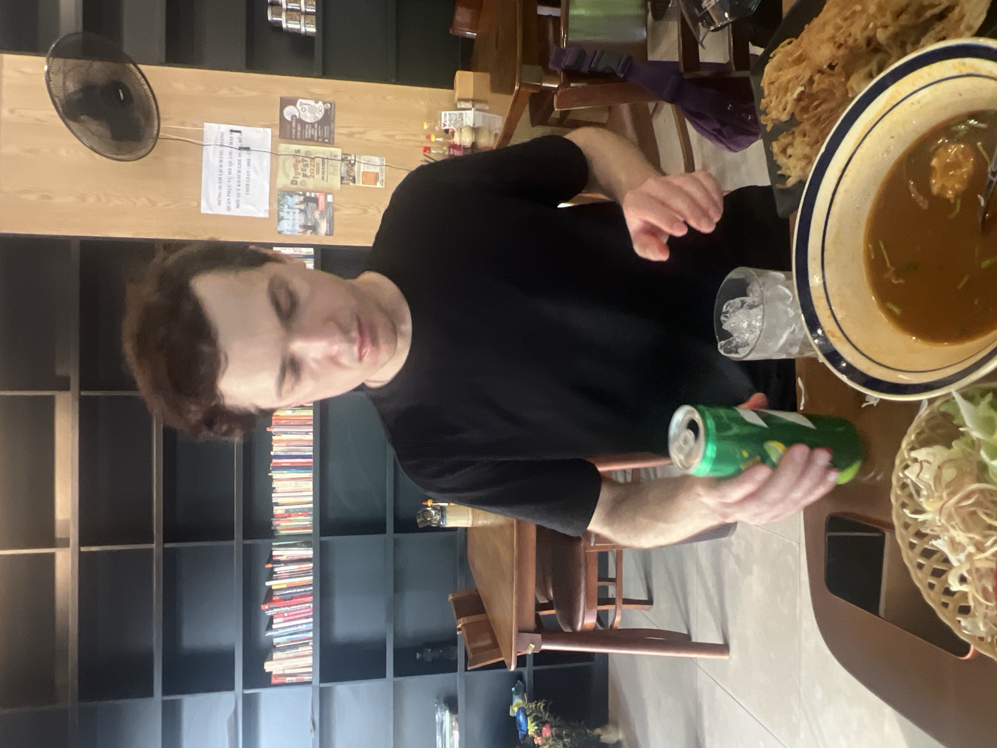 A man wearing a black t-shirt is seated at a wooden table in a restaurant. He is holding a green can of soda in his right hand and reaching for it with his left hand. In front of him, there is a glass of ice water, a bowl of soup, and a basket of food. The background of the image shows shelves filled with books and a wall-mounted fan. The man's expression suggests concentration as he is about to drink. The overall scene depicts a casual dining experience at a restaurant in Da Nang, Vietnam.