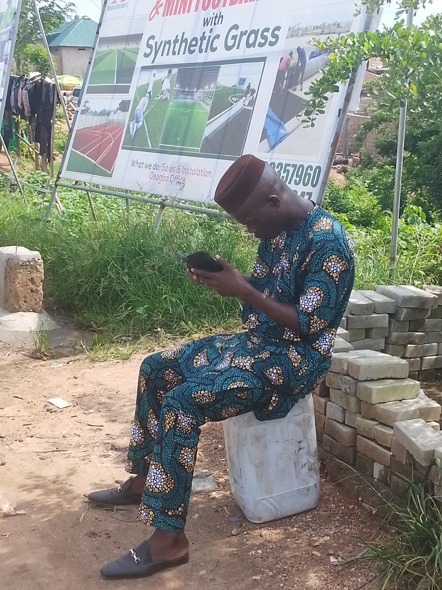 A Black man wearing a traditional blue and gold patterned outfit and a dark brown kufi cap is sitting on a white plastic container, looking down at a black smartphone he holds in his hands. He is outdoors, with lush green vegetation and a large billboard in the background. The billboard advertises "Mini Football with Synthetic Grass" and lists a phone number and "Oshogbo Office." To the right of the man, there is a stack of grey bricks. The ground is composed of dirt and small stones. The man's feet are clad in dark, slip-on loafers with gold buckles. The overall scene suggests an informal setting, possibly a roadside stall or a construction site, in Abeere, Nigeria.