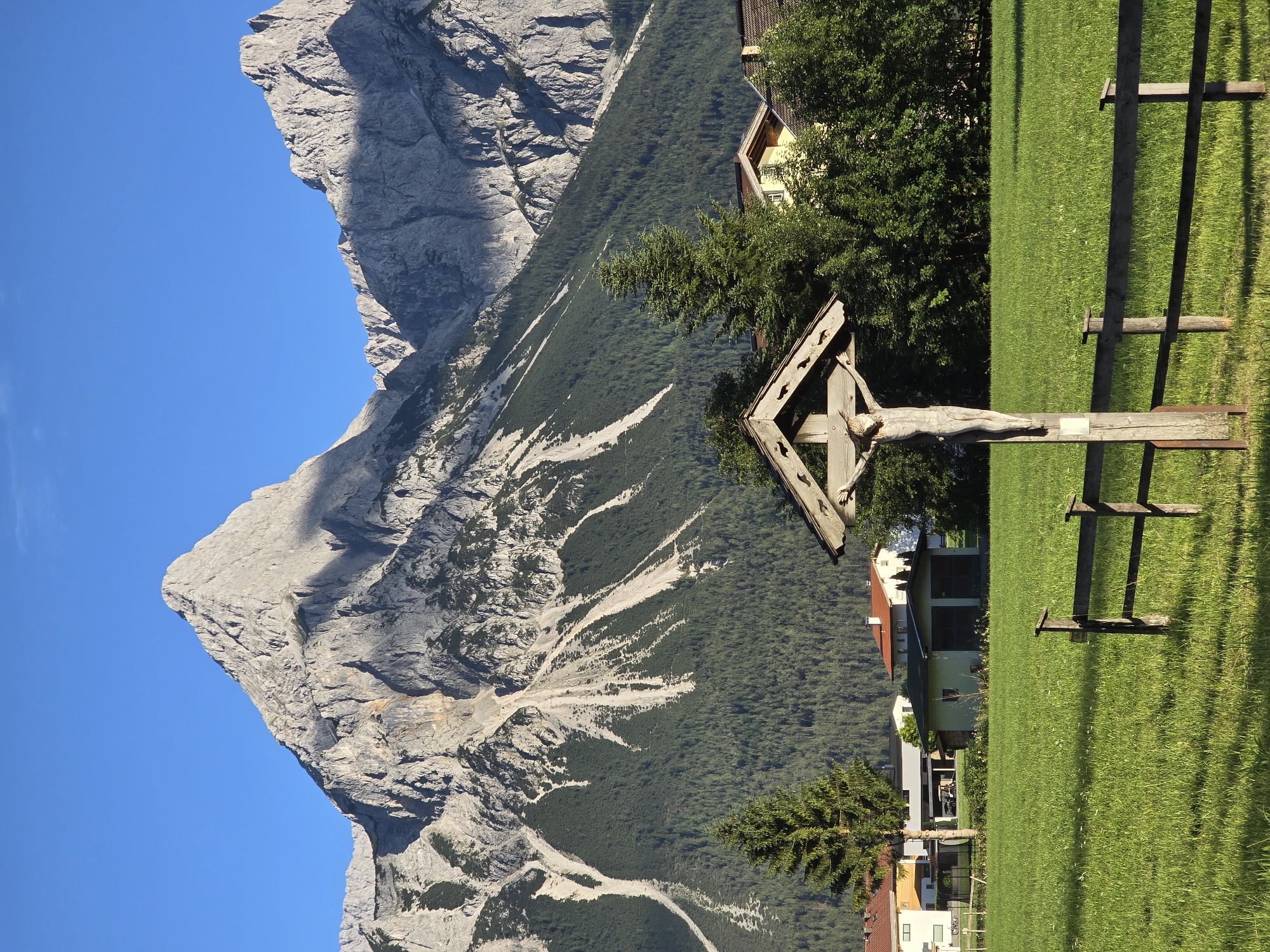 In Lermoos, Austria, a wooden wayside cross with a carved figure of Christ stands in a lush green meadow under a clear blue sky. The cross is positioned in the foreground, with a rustic wooden fence running horizontally across the meadow. In the midground, a cluster of trees and several buildings, including houses and a small garage, are visible. Behind the village, a majestic mountain range dominates the background. The mountains are rugged and imposing, with steep slopes covered in a mixture of rock and dense green forest. Streaks of light and shadow play across the mountain faces, highlighting their dramatic contours. The overall scene conveys a sense of tranquility and natural beauty, with the cultural element of the cross adding a spiritual dimension.