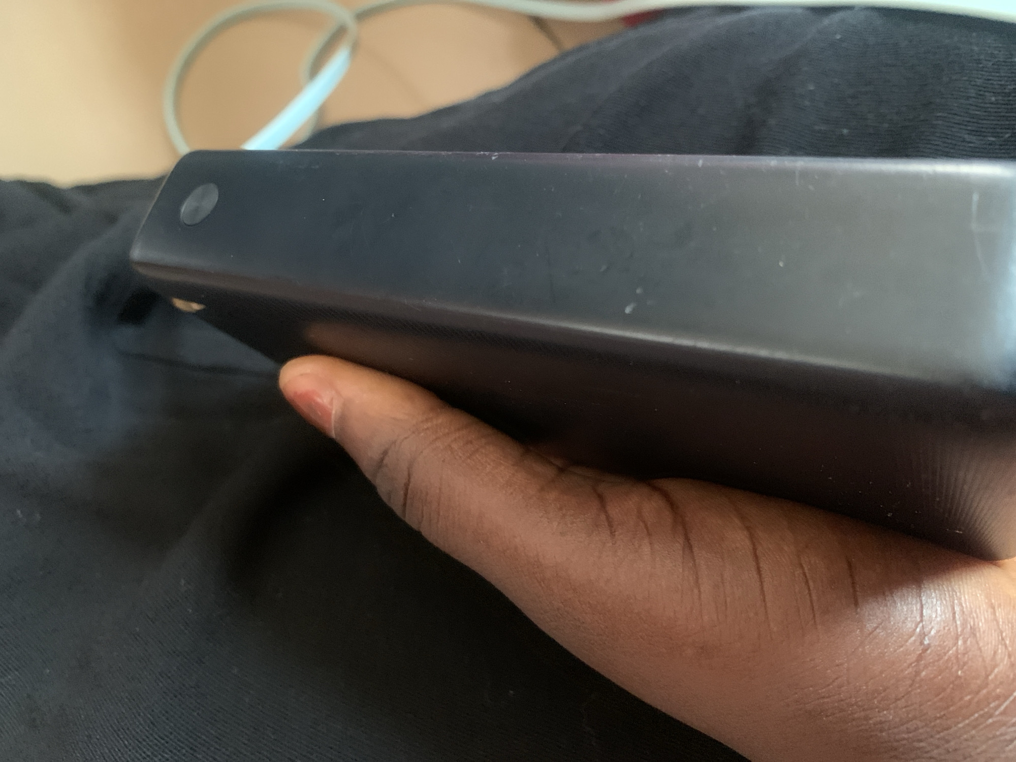 The image shows a close-up, low-angle shot of a person's hand holding a black electronic device. The hand is positioned on the right side of the frame, with the thumb and index finger wrapped around the device. The device appears to be rectangular with rounded edges, and a small circular button is visible on its surface. A light blue cable is partially visible in the background, extending to the right. The background consists of dark fabric, likely clothing, with some lighter, out-of-focus areas. The scene appears to be indoors, given the lighting and lack of natural elements. There is no discernible information about the specific location in Jauro Yinu, Nigeria, or any forensic details beyond the objects and their visible characteristics.