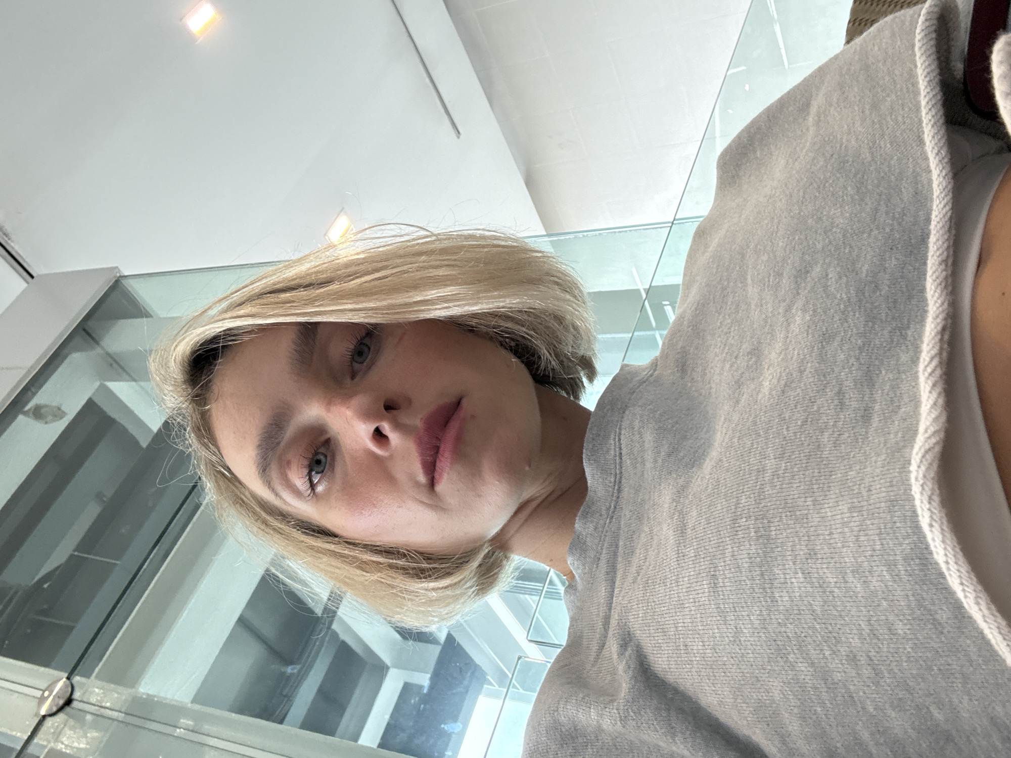 A blonde woman with light skin, blue eyes, and a slight smirk is looking up at the camera. She has on a grey sweatshirt and her hair is cut into a bob. The background is a blurry, modern interior with large windows and bright overhead lighting, suggesting an indoor public space such as an airport lounge or waiting area. The lighting is bright and diffuse, casting soft shadows on the woman's face and clothing. The scene is well-lit and the focus is on the woman's face, with the background out of focus. No specific location in Mai Khao, Thailand is identifiable from the visual elements of the image.