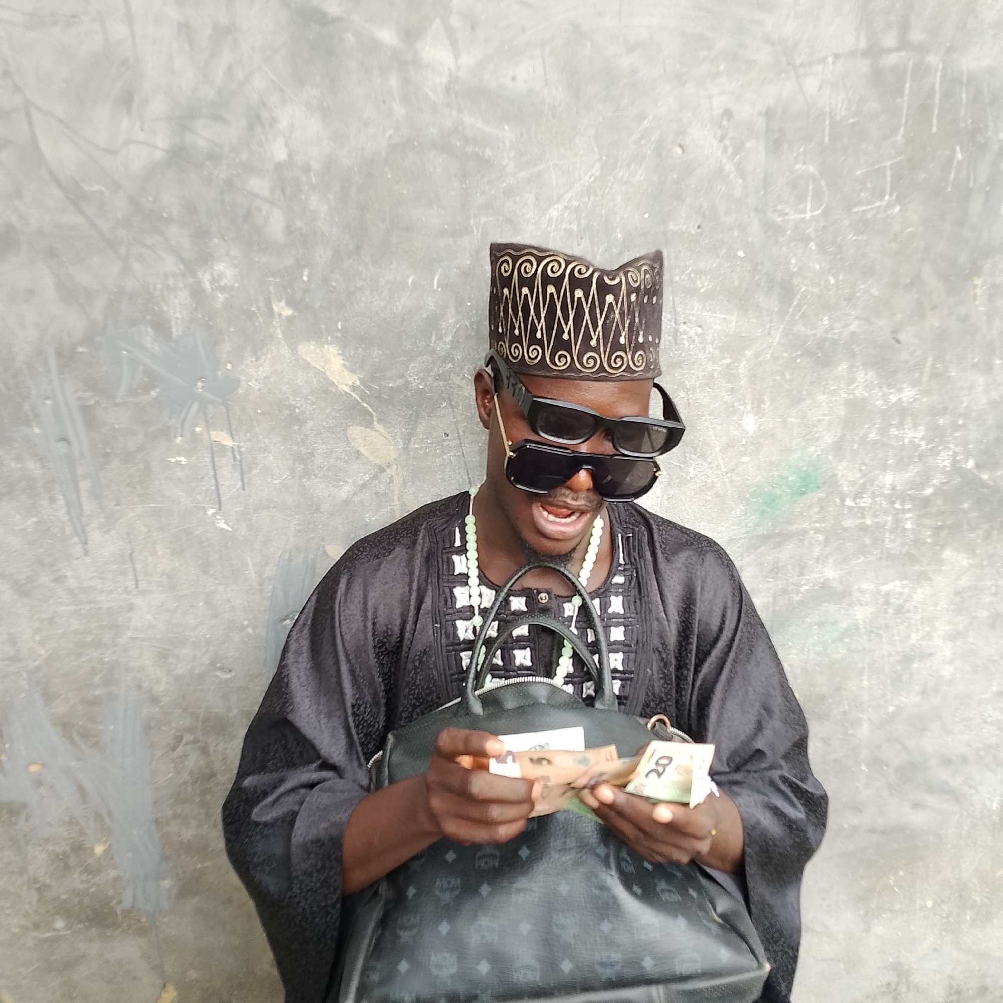 A man wearing a black traditional outfit, a patterned cap, and two pairs of black sunglasses, stands against a grey concrete wall. He is holding several banknotes in his hands, appearing to count them, in front of a large black handbag. The wall has some faint markings and splotches. The scene is shot from the chest up.