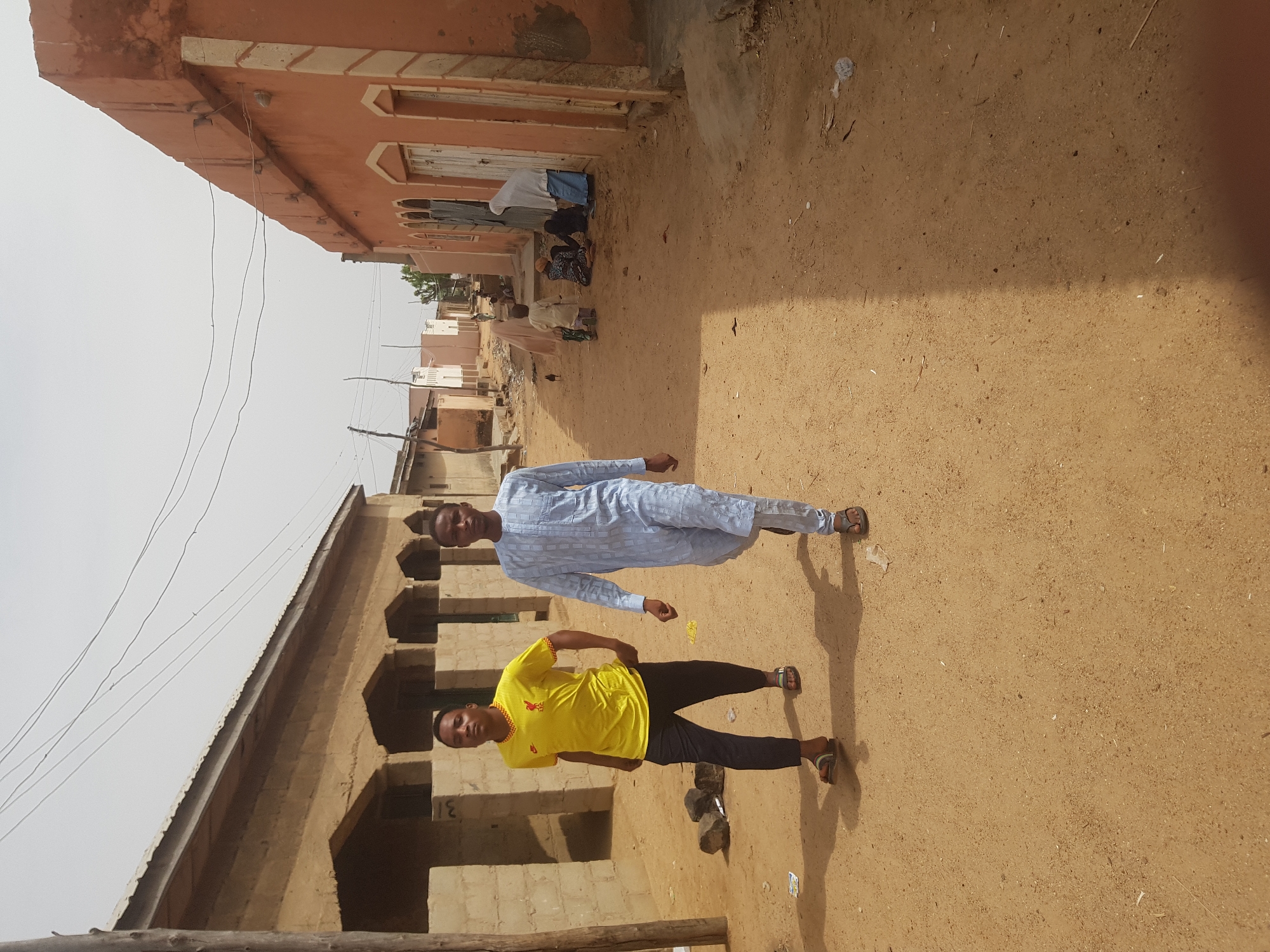 Two young men stand in a dusty, sunlit street in Dutsinma B, Nigeria. One wears a yellow t-shirt and dark pants, while the other is dressed in a light blue robe. The street is lined with buildings of earthen construction. In the distance, several individuals are seen praying near a building with peach-colored walls. Power lines extend across the sky.
