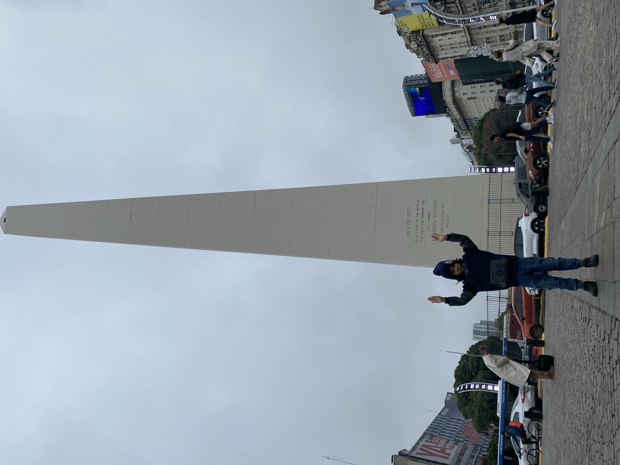 The image shows a tall, white, rectangular obelisk in the city of Buenos Aires, Argentina. A person in a blue jacket, jeans, and a blue hat is standing in front of the obelisk with their arms outstretched. The sky is cloudy. Behind the obelisk are buildings with digital billboards. Pedestrians and parked cars are visible on the street near the obelisk. The ground is paved with stone tiles.