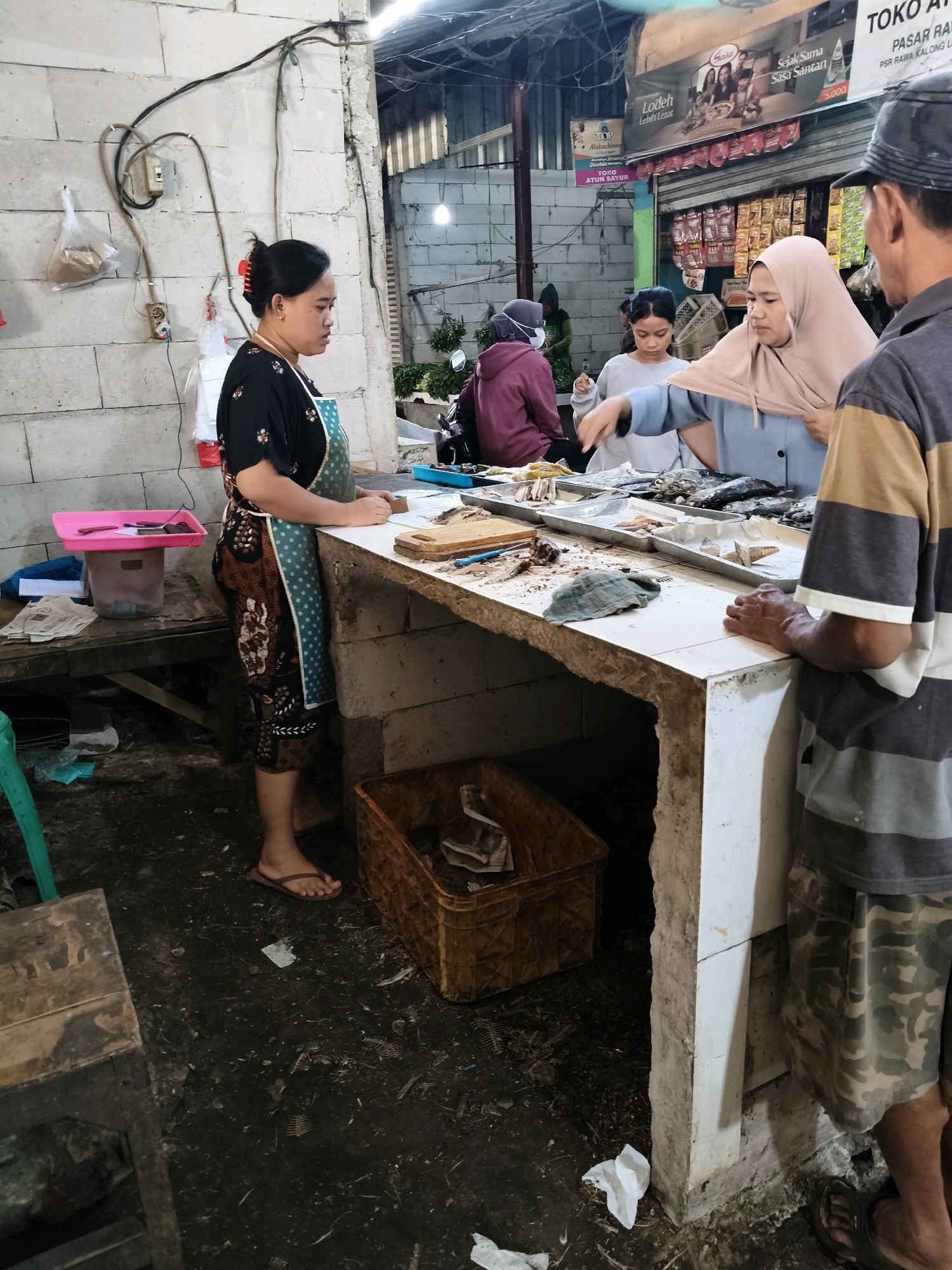 The image captures a scene inside a marketplace in Setiamekar, Indonesia. In the foreground, a woman wearing a black shirt, a patterned apron, and flip-flops stands behind a counter, appearing to be a vendor. She has her hair tied up with a red clip and is looking down. To her right, a woman wearing a light blue shirt and a hijab is interacting with a man in a gray and brown striped shirt and camouflage shorts, who is partially visible from the side. The counter is laden with fish, some of which are laid out on trays. Behind the counter, there are shelves stocked with various goods. The environment appears somewhat cluttered and utilitarian, with a concrete wall visible in the background and a basket on the floor. The lighting suggests an indoor market setting, possibly with overhead lights illuminating the area. Several other individuals, including one wearing a headscarf, are visible in the background, contributing to the bustling atmosphere of the marketplace.