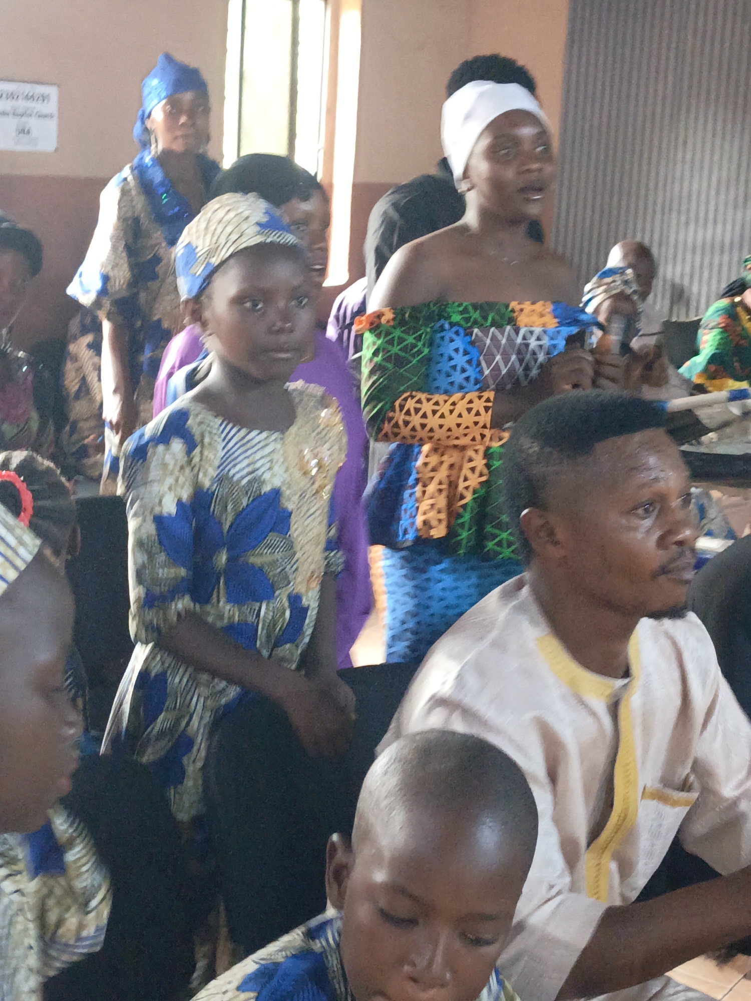 Akwanga, Nigeria. A group of people, including adults and children, are inside a building, likely a church or community center. Some individuals are wearing traditional African clothing with colorful patterns and designs. A woman with a white headband holds a microphone and appears to be speaking or singing. A man in a light-colored shirt with yellow trim is visible in the foreground. The environment is indoors with a window in the background, and a sign is visible on the wall.