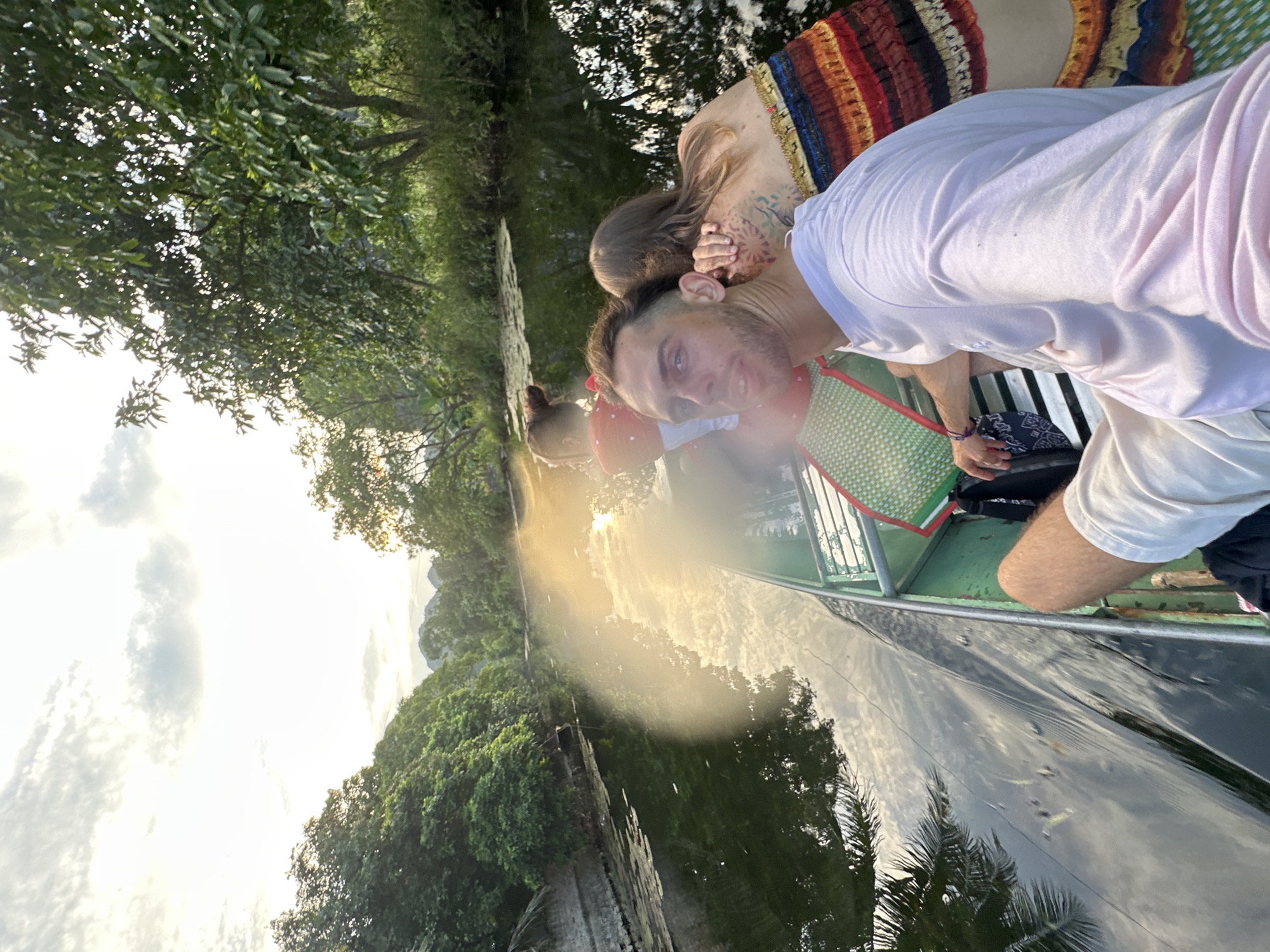 A selfie is taken from the front of a long, narrow boat moving down a calm waterway lined with lush green vegetation. The sky above is bright with clouds, and the sun is setting or rising, casting a warm glow on the water. Three individuals are visible in the boat. In the foreground, a young man with short, dark hair and a beard looks directly at the camera, wearing a light pink t-shirt. To his right and slightly behind him, a person with long, light brown hair has their arm around the man's neck and is facing away from the camera. Their back is adorned with intricate tattoos. Further back in the boat, a young child wearing a red shirt with white polka dots is also visible, facing away from the camera. The boat is green and has seating with colorful mats. The environment suggests a natural setting, likely a river or canal, with dense trees and foliage on either side of the water. The overall impression is one of a leisurely boat ride in a scenic natural landscape. The location is Nam Hoa Lư Ward, Vietnam.