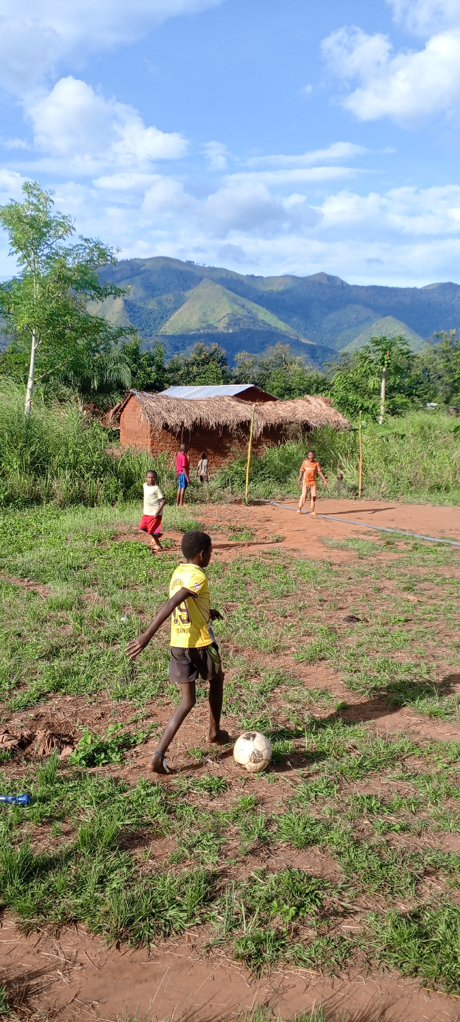 The photograph depicts a group of children playing soccer in what appears to be Ngalula, Democratic Republic of the Congo. A boy in a yellow soccer jersey and black shorts is dribbling a worn soccer ball in the foreground. Behind him, other children are positioned on a dirt field, one wearing a red top and red shorts, and another in an orange shirt. In the background is a simple, mud-walled dwelling with a thatched roof. Beyond the house is a landscape of rolling hills and mountains under a partly cloudy sky. Tall grass and vegetation are present.