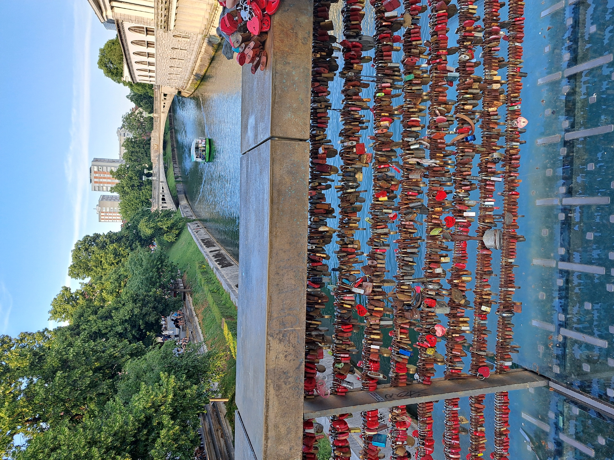 The image depicts a wide, calm river flowing through a city under a clear blue sky. A pedestrian bridge, adorned with numerous colorful love locks, spans across the river in the foreground. The locks are attached to the bridge's railings, creating a dense tapestry of red, gold, silver, and blue. Below the bridge, the river's surface reflects the sky and the surrounding architecture. A small, green boat filled with passengers navigates the water in the mid-ground, heading towards a decorative stone bridge further down the river. The riverbanks are lined with lush green trees and stone embankments. On the right bank, an ornate building with arched windows and a stone facade stands prominently. On the left bank, a grassy slope is visible with people seated and walking along a pathway. In the background, modern high-rise buildings rise above the trees, indicating an urban setting. The overall scene conveys a sense of urban tranquility and romantic tradition.