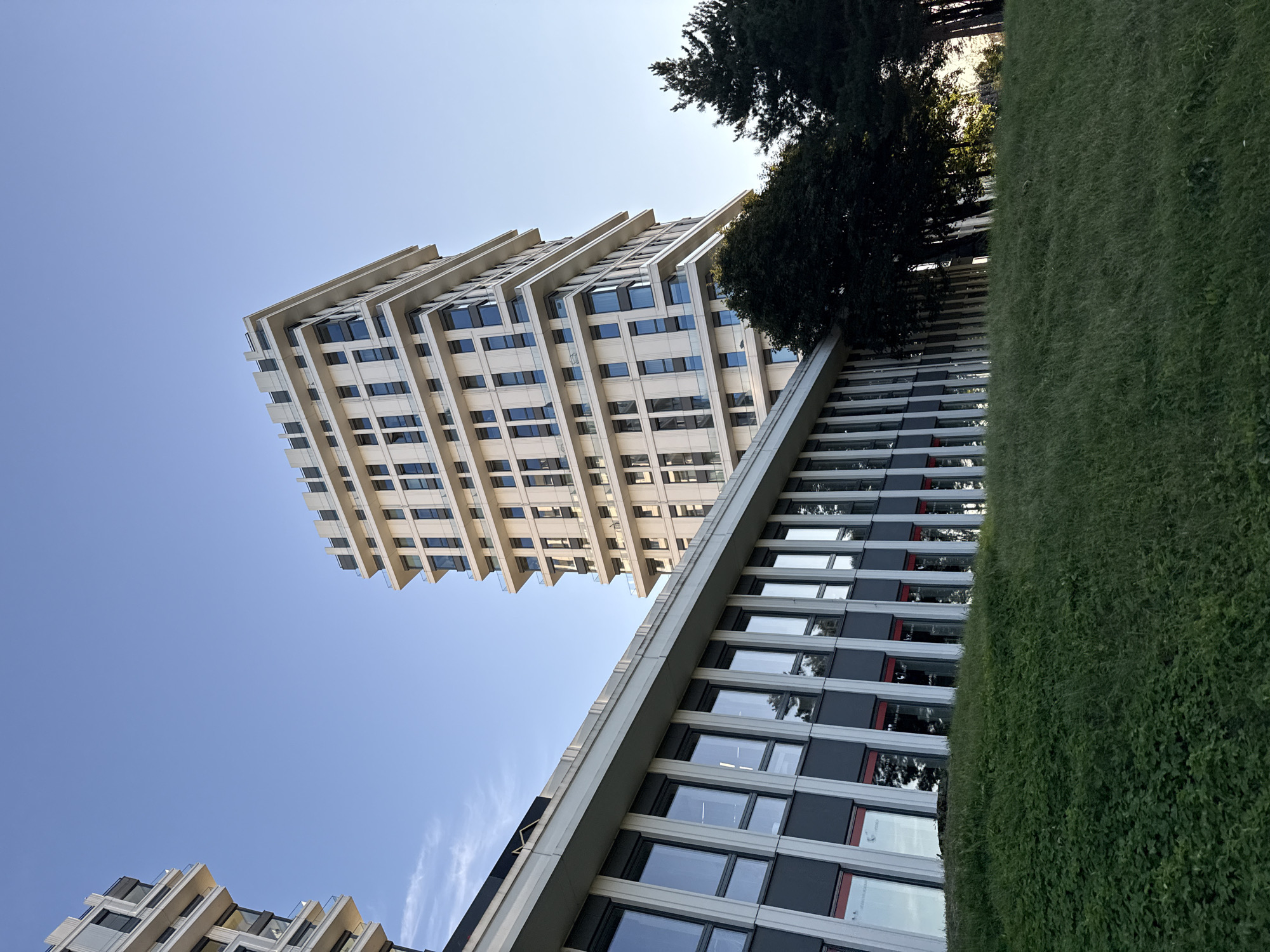 This is an exterior, eye-level, full shot of modern buildings in Tallinn, Estonia, on a clear day with a blue sky. The main subject is a tall, multi-story building with a white facade, featuring numerous windows and balconies. To the left and foreground is a lower, horizontal building with a gray and white facade and a long row of windows. The buildings are situated on a grassy slope, with dark green trees visible to the right. The lighting suggests it is late afternoon or early evening, with sunlight illuminating the side of the tall building. There are no people visible in the scene.