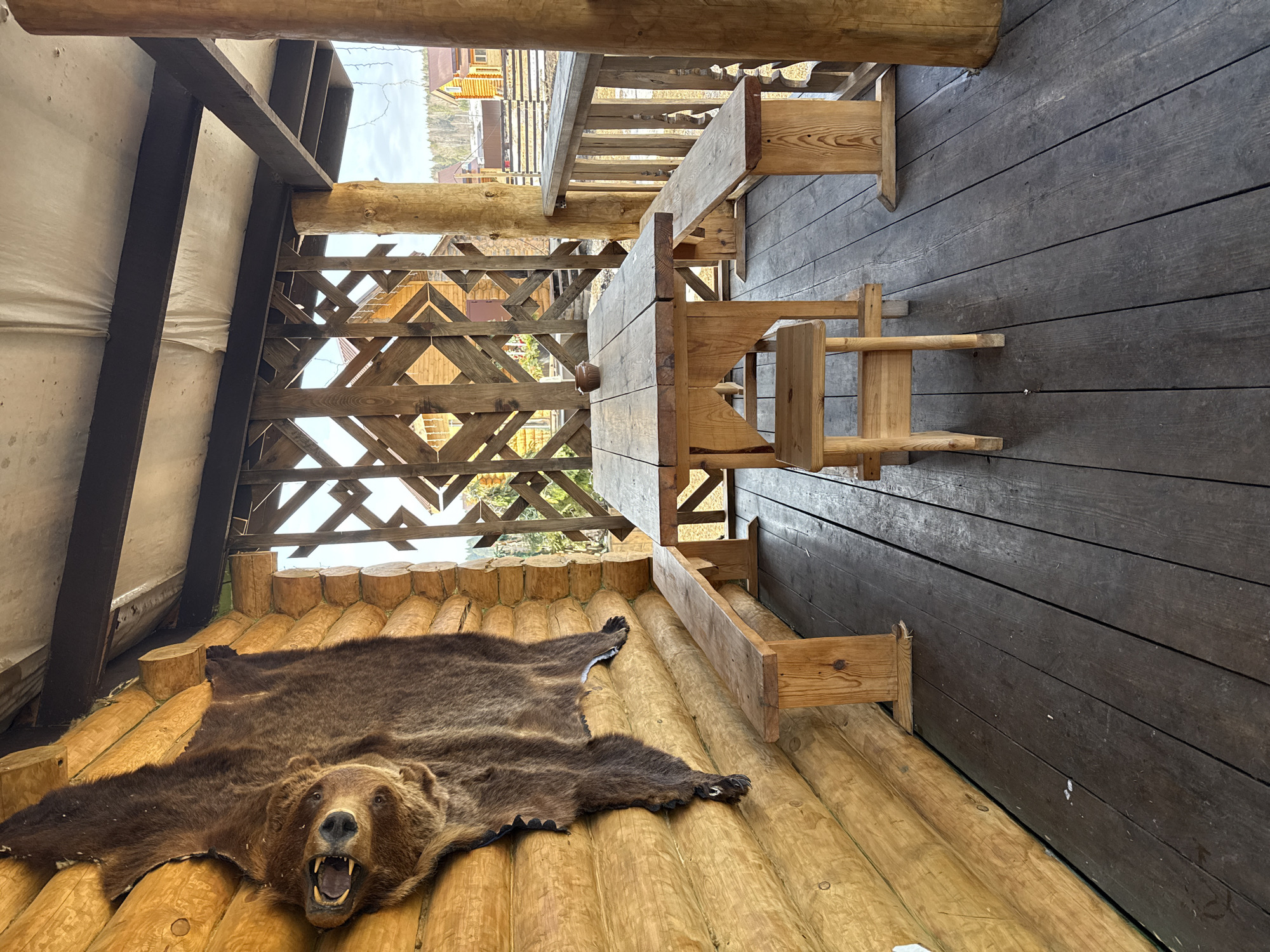 The image depicts an unoccupied rustic porch or veranda in Tyulyuk, Russia. A large, dark brown bear hide is mounted vertically on the left log wall, featuring the bear's head with an open mouth displaying teeth. The porch floor consists of dark, weathered wooden planks. In the center, a heavy, light-colored wooden table is present, flanked by two matching wooden benches. A smaller, low wooden stool is positioned at the near end of the table. A single small, light brown pottery bowl rests in the center of the tabletop. The ceiling features dark support beams and a lighter, possibly fabric or tarp-like material. A decorative wooden lattice screen with diamond patterns forms a partial wall in the background, allowing views of other wooden structures, a fence, distant trees, and a cloudy sky beyond. Large, raw wooden posts support the porch structure. No individuals are present.