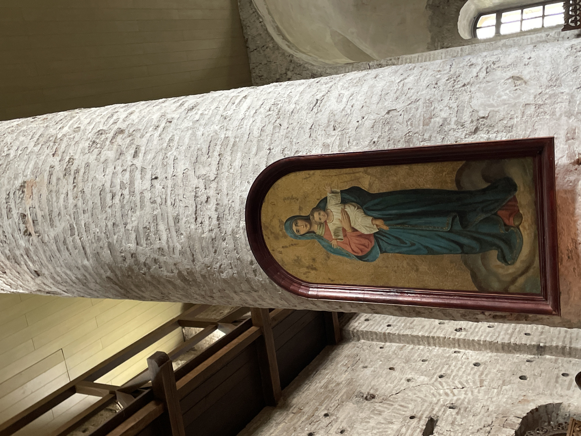 The media file captures an interior view of a religious building, likely a church or monastery, in Hrodna, Belarus. A large, textured, light-colored pillar, possibly whitewashed stone or plaster, dominates the left side of the frame. Embedded within an arched recess on the right side of this pillar is a framed icon depicting the Virgin Mary holding the infant Jesus.
The icon portrays Mary in blue and red robes, gazing forward with a solemn expression, cradling Jesus, who is dressed in white and raises one small hand. The frame of the icon is dark wood.
To the right of the pillar, an old stone and brick wall is visible, featuring a small, arched window with grilles in the upper right, allowing natural light into the space. Further down the wall, faint holes are scattered across its surface. In the lower-left background, wooden beams, planks, and a railing suggest an upper gallery or part of a ceiling structure. The overall lighting indicates daytime. There is no discernible activity taking place, and no visible text is present in the image. The atmosphere is one of historical reverence.