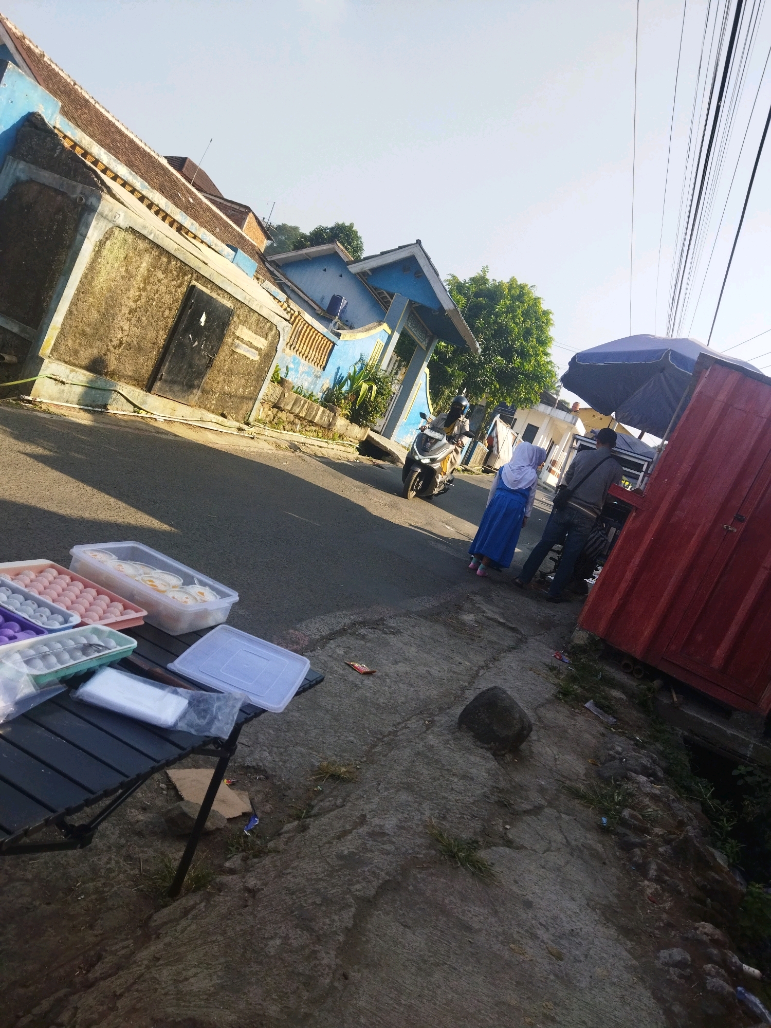 The image depicts an outdoor scene on a sunny day, likely in Sudajaya Girang, Indonesia, based on the provided context. A street runs diagonally across the frame, with a weathered building on the left and a red structure resembling a shipping container on the right. In the foreground, a black folding table is laden with various plastic containers filled with food items, possibly eggs and desserts. Some of these containers are arranged in rows, and one is a large rectangular container with a lid.
On the street, a person on a motorcycle is visible, heading away from the viewer. Further down the street, near the red structure, two individuals are interacting. One, wearing a blue hijab and dress, appears to be a young woman or girl, while the other is an adult male in a gray jacket and dark pants. They are standing under a large blue umbrella, suggesting a small market stall or roadside business. The red structure on the right may be part of this stall.
The environment includes a concrete sidewalk next to the street, with patches of grass and debris. Overhead, power lines stretch across the sky, and trees can be seen in the background, indicating a semi-urban or rural setting. The overall impression is that of a casual, everyday street scene with some form of commerce taking place.
