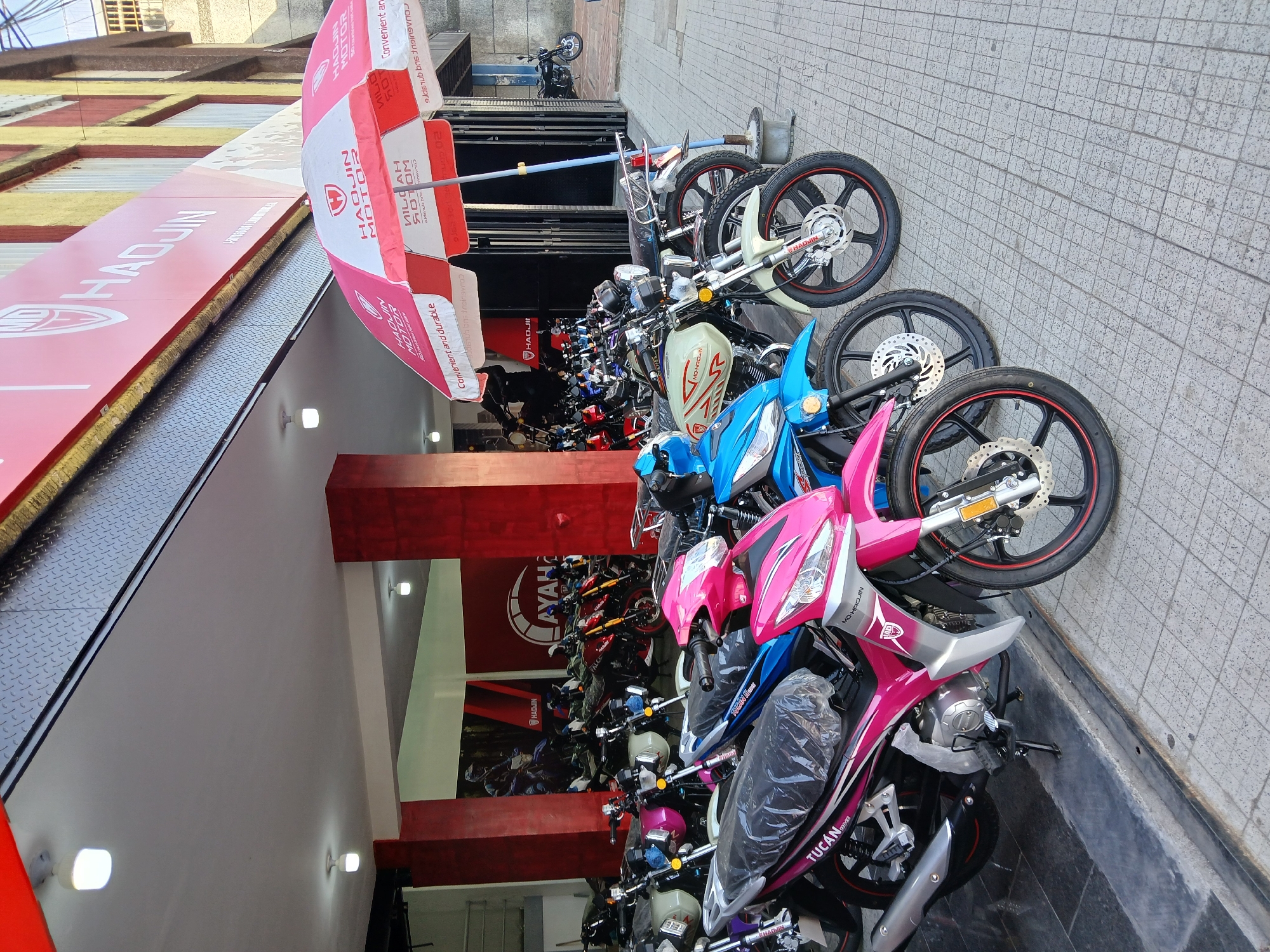 The image depicts a storefront for "Haodjin Motor" in Ciudad Guayana, Venezuela. The establishment is a motorcycle dealership or shop, indicated by the numerous new motorcycles lined up on the sidewalk in front of the store. The motorcycles vary in color, including pink, blue, green, and white. A large red-and-white "Hadjin Motor" branded umbrella is set up outside. The storefront features red signage with the "Hadjin Motor" logo and branding. The sidewalk is paved with gray tiles.