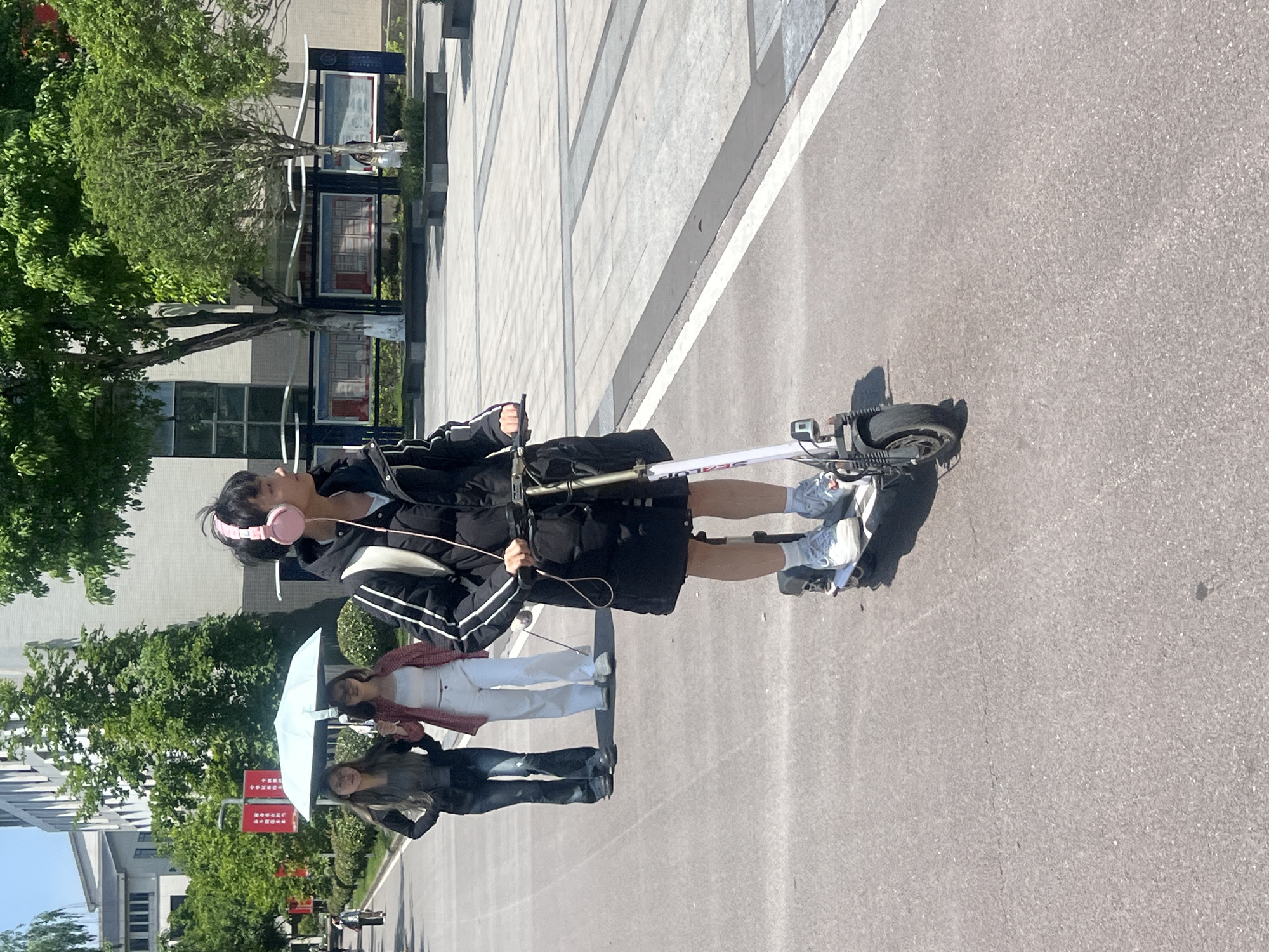 The image captures a sunlit outdoor scene in Jiangjin, China, showing three young individuals on a paved urban path or campus area.
The central subject is a young person with short dark hair, wearing pink over-ear headphones, a black jacket with white arm stripes, a black skirt or shorts, white socks, and light-colored sneakers. They are standing on a white and black electric scooter, holding its handlebars. The scooter's stem clearly displays "SEALUP". The person appears calm, looking slightly upwards.
Behind and to the left of the main subject are two young women. The closer woman holds a white umbrella, wearing a white top, light-colored pants, and a reddish-brown plaid shirt unbuttoned over it. She has long dark hair and a faint smile. Further back, another woman with long dark hair is seen wearing a dark jacket and blue jeans, also with a subtle smile.
The setting features a wide, light grey asphalt path on the right, bordered by a white line, and a patterned light grey concrete paved area with dark grey stripes on the left. In the background, modern multi-story buildings with glass windows are visible, surrounded by lush green trees and bushes. Several red signs with Chinese characters are posted on poles among the trees, serving as information boards or traffic signs.
The bright sunlight casts distinct shadows, indicating it is likely midday or early afternoon on a clear day. The overall atmosphere appears casual and relaxed.