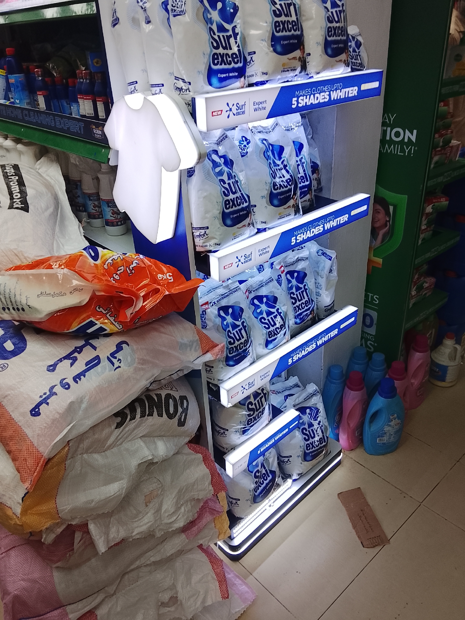 This indoor scene shows a display of laundry detergent in a store, likely in Gulgasht Colony, Pakistan, given the text on the packaging. Stacks of large white bags with Arabic script and the word "BONUS" printed on them are on the left side. An orange bag is placed on top of the white bags. To the right, a shelf displays "Surf Excel" laundry detergent in smaller, individual packages. The shelf has LED lights outlining each level, and blue signs reading "MAKES CLOTHES UP TO 5 SHADES WHITER." Above the detergent shelf, other cleaning products are visible, along with a white illuminated t-shirt shape. To the right of the detergent shelf, laundry detergent bottles in pink and blue colors are seen. The floor is made of light-colored tiles, and a piece of cardboard is lying on the floor.