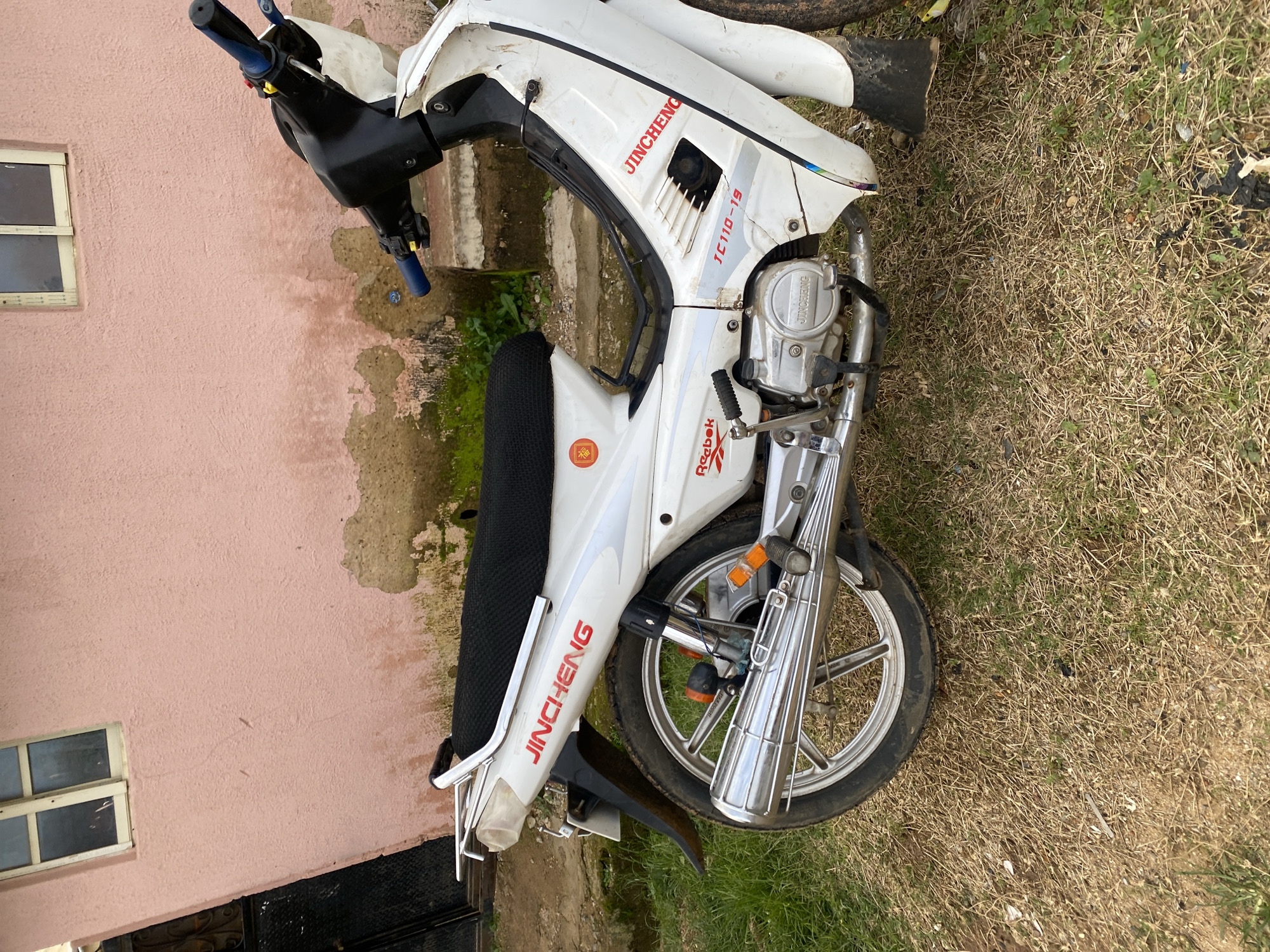 The image shows a white Jincheng motorcycle parked on dry, grassy ground in front of a pink, weathered building. The motorcycle has red "JINCHENG" lettering on its side and a black seat. The building appears to be a residential structure with a window and a dark metal door. The environment suggests an outdoor setting in a possibly rural or less developed area, consistent with the location provided. There are no people visible in the image, and the motorcycle is stationary. The overall scene is a simple depiction of a parked vehicle against a backdrop of a building and natural elements.