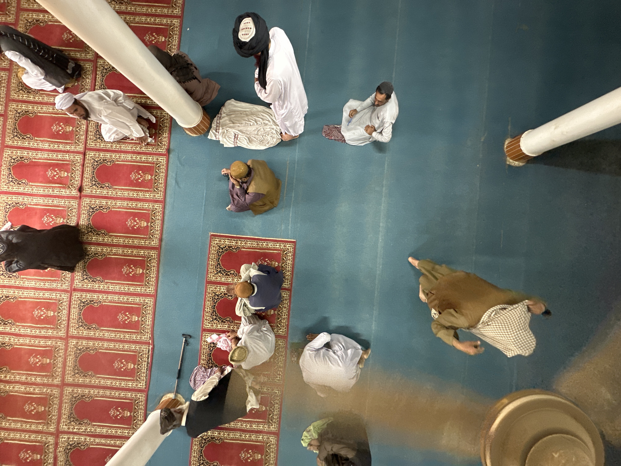 This is an aerial, high-angle, full shot of a mosque interior where several men are gathered on prayer mats. The floor is covered with a blue carpet, and a series of red prayer rugs with intricate gold and black patterns are laid out in rows. In the upper section of the image, several men are dressed in white robes and turbans, some are seated in prayer, others are standing. A large white cylindrical object, possibly part of the mosque's architecture or a fan, is diagonally placed across the scene. In the lower left, a man in a brown robe and white checkered skirt is standing and pointing upwards. Further down and to the right, another man in a white robe and patterned headscarf is seated. The overall atmosphere suggests a religious gathering or prayer service. The context of Kanozai, Pakistan is provided externally and is not visually discernible from the image alone.