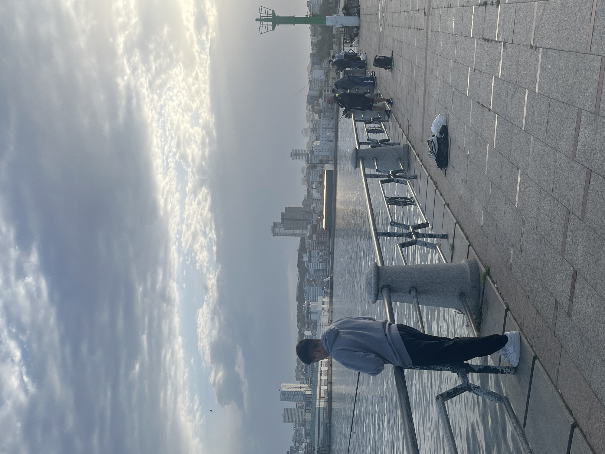 The image shows a man fishing from a quay or boardwalk alongside water. He is wearing a grey hoodie, dark trousers, and white sneakers. He is leaning on a metal railing, looking down at the water. There are other people fishing further down the quay. In the background, there is a city skyline with buildings of varying heights, and a green and white structure that resembles a lighthouse. The sky is overcast with various cloud formations. The quay is made of grey paving stones. The location is likely in A Coruña, Spain, due to the presence of a recognizable structure that is consistent with the city's lighthouse and waterfront area. The water appears calm, and there is a reflection of sunlight on its surface.
