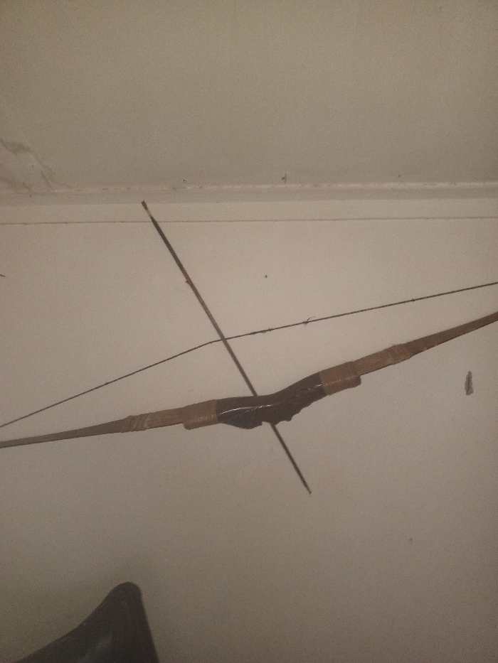 A bow and arrow hang against a white wall. The bow, made of wood, is dark in the center with lighter brown tips. The string stretches tautly between the bow's ends. An arrow intersects the bow's center, pointing downwards. A dark, indistinct object is partially visible in the lower left corner. The wall has minor imperfections near the ceiling.