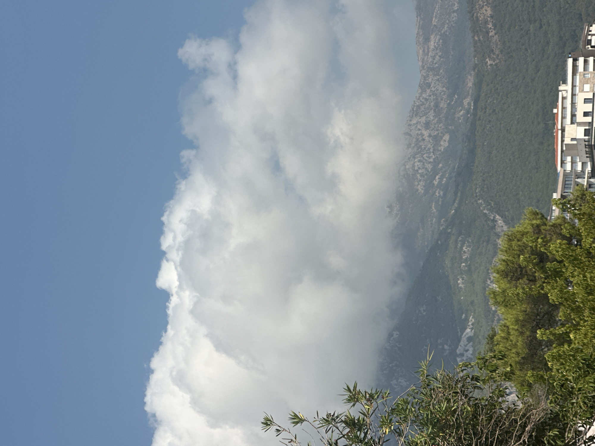 The image depicts a scenic view of the coastline in Boreti, Montenegro. In the foreground, lush green trees dominate the lower part of the frame, with branches extending into the upper left corner. Behind the trees, a modern, multi-story building with white balconies and a red-tiled roof is visible.
The midground features a sloping mountain range covered in dense green foliage, interspersed with rocky outcrops. The higher elevations of the mountains are shrouded in a thick blanket of white clouds, which also extend into the upper portion of the image, obscuring the sky in places. The sky itself is a clear, vibrant blue, particularly noticeable in the top right corner. The overall impression is one of a natural landscape meeting urban development, under a bright, partially cloudy sky.