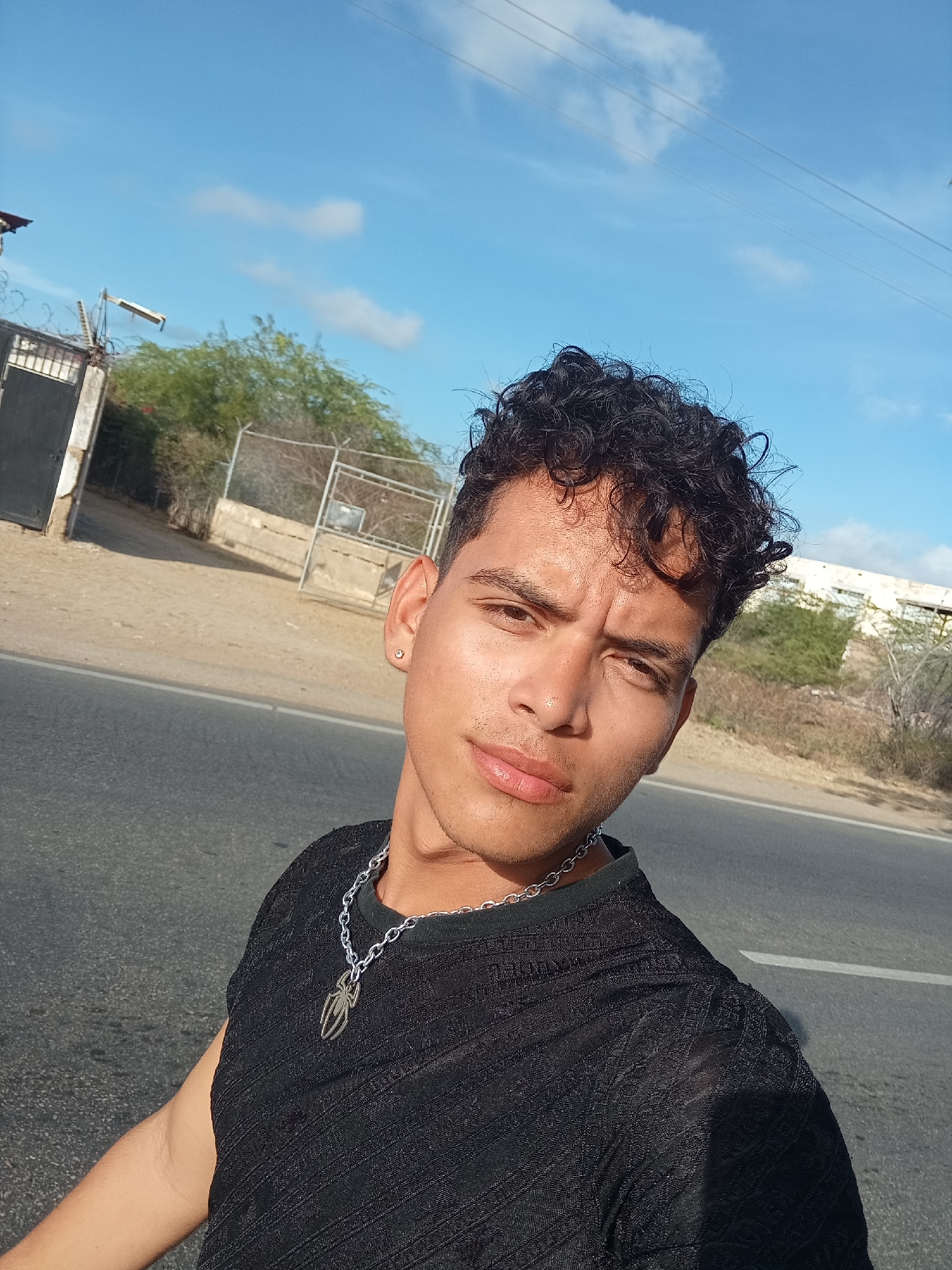 A young man takes a selfie outdoors on a bright, sunny day in Juan Griego, Venezuela. He is positioned in the foreground, facing the camera directly, with a serious or slightly intense expression, his eyebrows slightly furrowed. He has short, dark, curly hair and a light brown complexion. He wears a black, textured short-sleeved shirt and a silver-colored chain necklace with a distinctive spider-shaped pendant. A small, sparkling stud earring is visible in his left ear.
The background shows a semi-arid landscape with sparse, dry-looking trees and bushes under a clear blue sky with a few wispy clouds. A paved road, identifiable by a faded white line, runs horizontally behind him. To the left, there's a dark metal gate with barbed wire on top, and a chain-link fence extends into the distance. Power lines are visible against the sky, particularly in the upper right. Further back, on the right side of the image, parts of a light-colored building can be seen on a slightly elevated area. The strong sunlight suggests it's daytime, possibly late morning or afternoon. There is no visible text in the image.