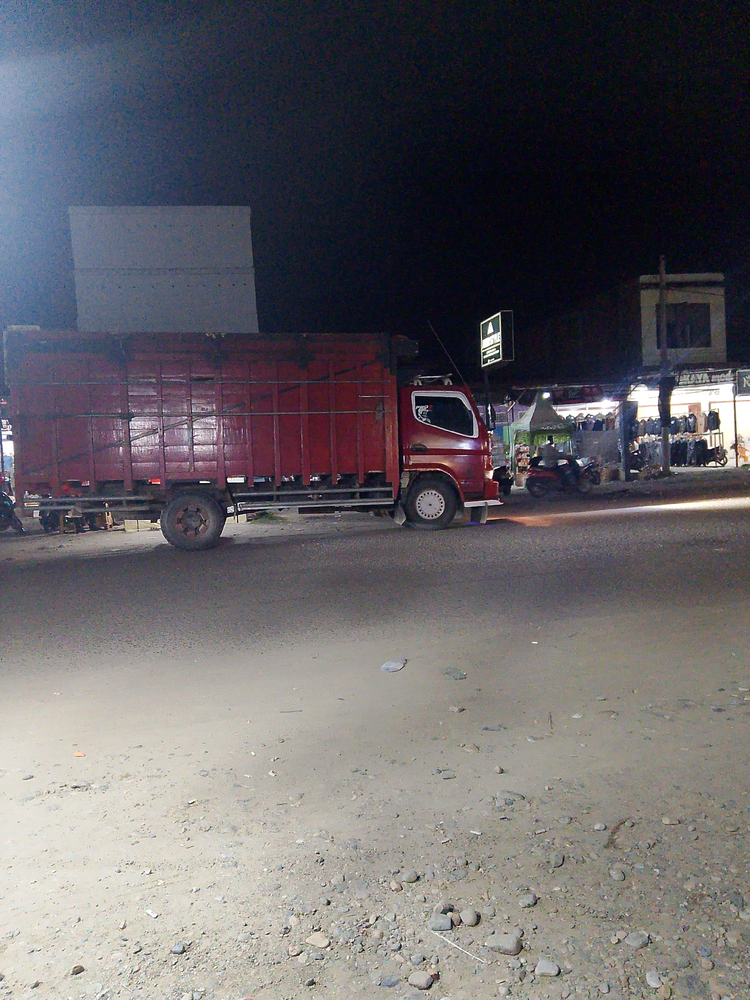 In Gampongpaya, Indonesia, a dimly lit scene unfolds at night. A large, red truck, its side adorned with horizontal slatted panels, is parked on a dirt and gravel road. The truck's tires are visible, and its cabin lights appear to be on. To the right of the truck, a row of stalls or shops line the street, illuminated by artificial lights. Some of these stalls display clothing on racks. Several motorcycles are parked in front of the stalls, and a few individuals are visible in the background, interacting near the shops. A prominent green sign with white text, possibly indicating a business, hangs above one of the stalls. The background is dark, indicating the nighttime setting, with some buildings and structures visible. The ground in the foreground is rough and uneven, composed of dirt and small stones.