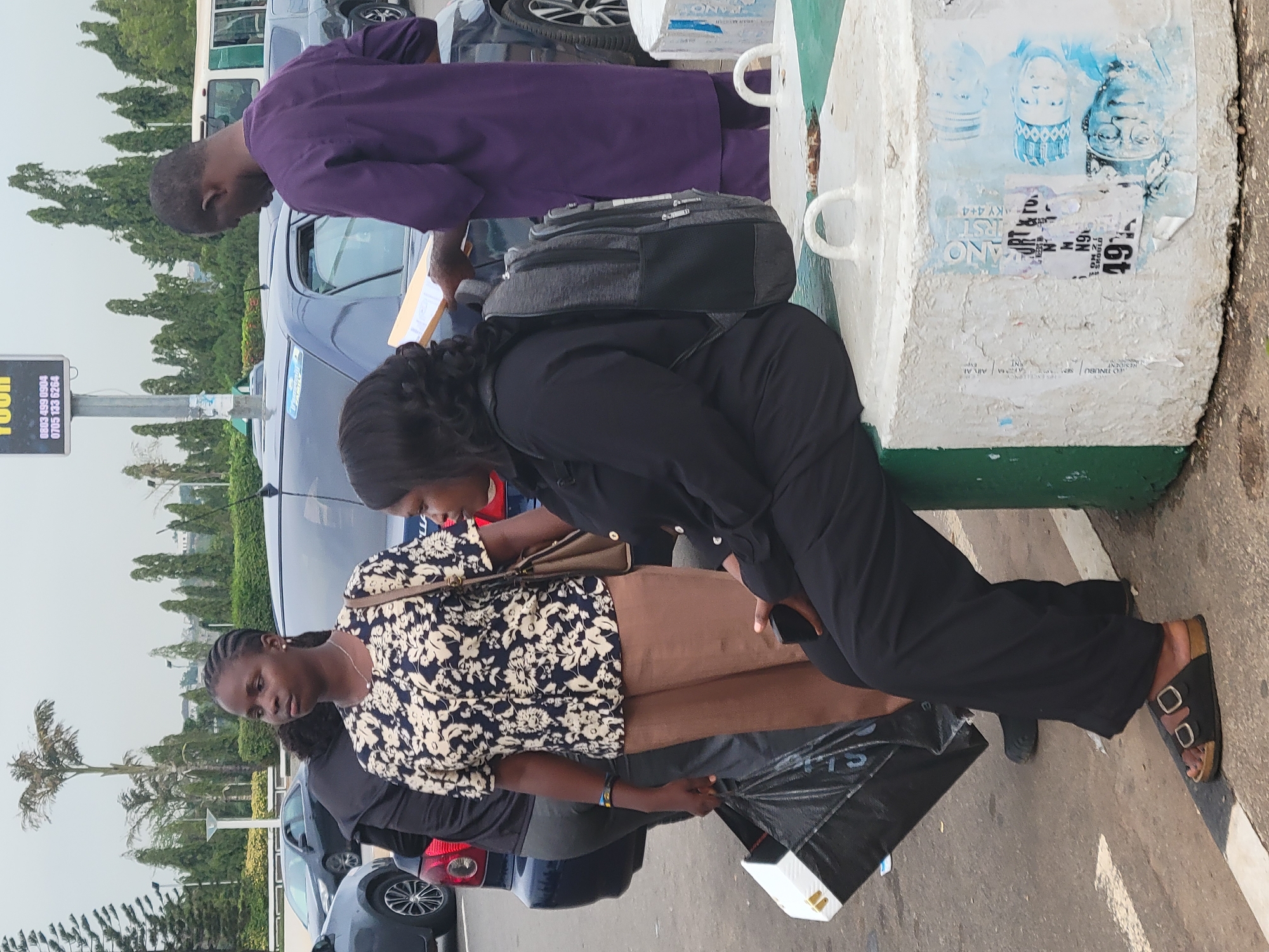 The image, originally rotated 90 degrees clockwise, presents an outdoor scene in Abuja, Nigeria, during daytime, likely under an overcast sky. Three individuals are gathered near parked cars and a concrete barrier.
In the foreground, a woman in a black and white floral patterned top and brown pants holds a black plastic bag, possibly branded "SLIP" or "SPORTS." She has braided hair, wears a wristband, and a brown shoulder bag. Adjacent to her, another woman in a black top and pants leans forward towards a concrete barrier, wearing a grey backpack and sandals. Her face is partially visible. Behind them, a man in a purple tunic stands by a dark blue car, looking down at a white envelope or document he holds.
Several cars are parked in the background, alongside green trees and distant buildings, characteristic of an urban environment. The large concrete barrier, painted white with a green base, features metal handles and is adorned with various torn posters and papers. Visible text includes "49," "6N," "ANO," "IRST," and "KY 4+4," with fragments like "All Al" and "TINUBU" also discernible. A prominent poster on the barrier displays images of three men, likely political figures. A pole nearby holds a sign with "FOOD" partially visible at the top, along with two phone numbers: "0803 499 0904" and "0705 133 6264."
The individuals appear to be standing or waiting, with neutral to focused expressions, in what seems to be a casual public gathering or transit area.
