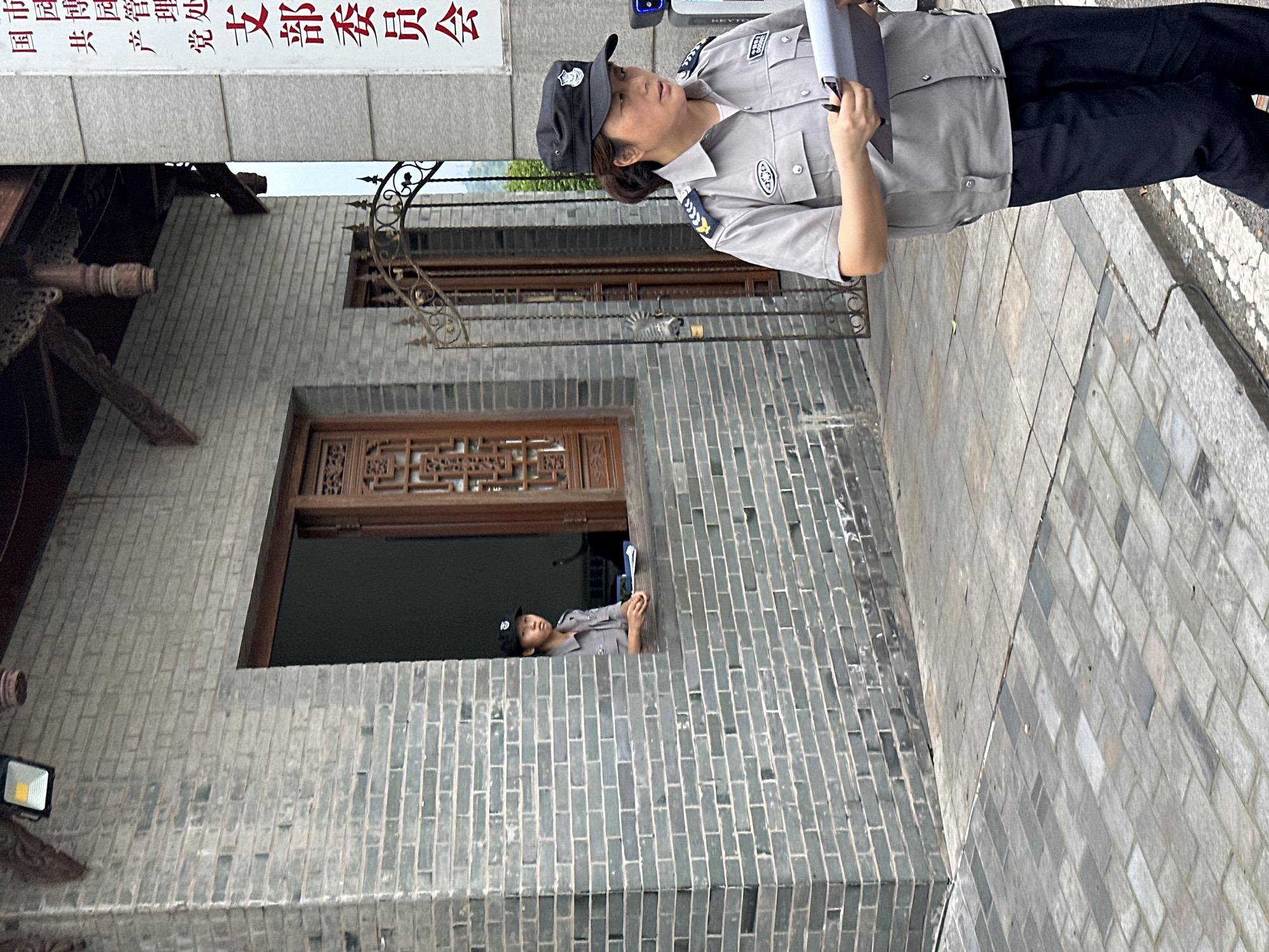 The photograph shows a uniformed security officer standing on a sidewalk adjacent to a building in what is reported to be Liangjiang, China. The officer is holding a clipboard and is looking up and to the left. A second uniformed security officer is visible in a window of the building. The building is constructed of grey brick, has a decorative window frame, and has a sign written in Chinese characters above the window. The sidewalk is paved with grey stones.