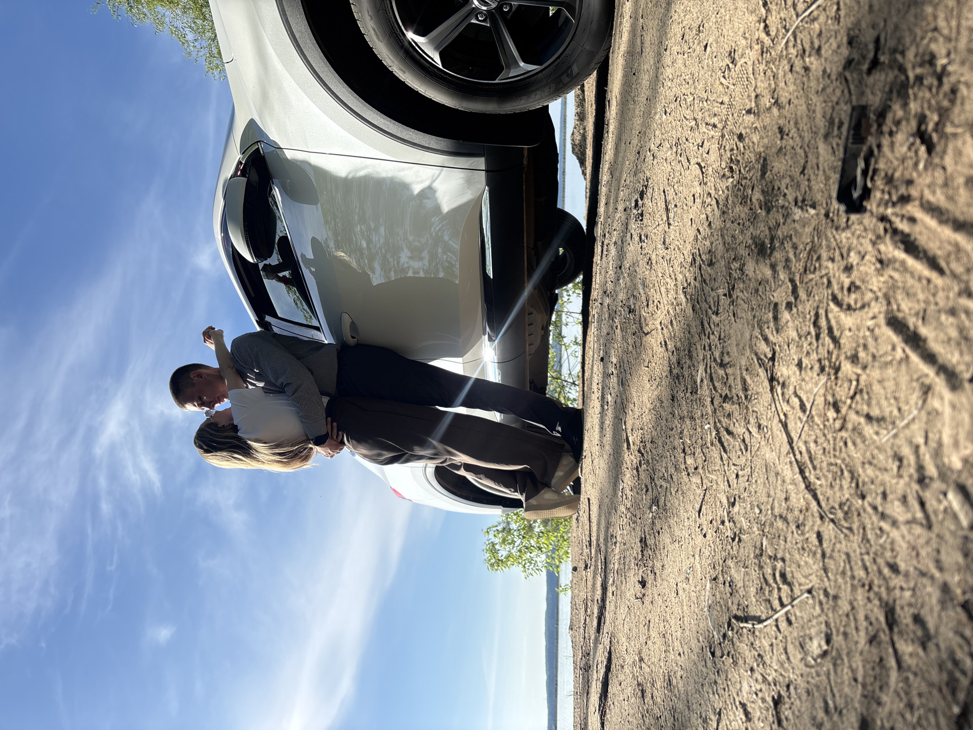 A couple is embracing beside a white car. The location appears to be a dirt road near a body of water under a blue sky with wispy clouds. The image orientation is rotated, placing the sky and the couple's feet on opposing sides of the shot. It's impossible to confirm the city of Chaikovsky, Russia, based solely on the provided image.