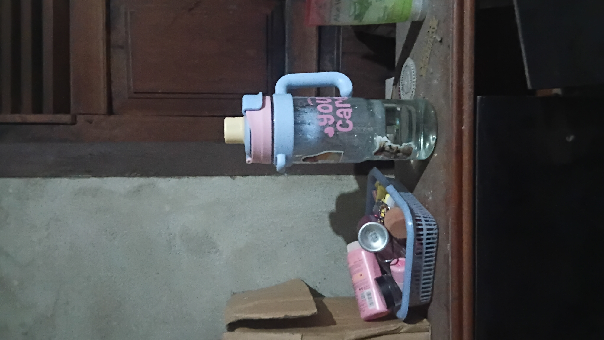The image depicts a still life scene on a wooden surface, possibly a shelf or dresser, in a dimly lit room. On the left, a blue plastic basket contains various items, including a pink lotion bottle, a silver cap, makeup sponges, and other small objects. To the right of the basket is a clear plastic water bottle with a blue handle and lid, decorated with stickers of cats and pink text that reads "you car". The bottle contains a clear liquid. Further to the right, partially visible, is a green plastic bottle, possibly a personal care product. The background consists of a textured gray wall on the left and a dark wooden door with horizontal slats on the right. The overall environment appears to be domestic, with a slightly cluttered and informal arrangement of personal items. The image does not contain any information that would place it in Prabumulih, Indonesia, or suggest any specific actions or interactions.