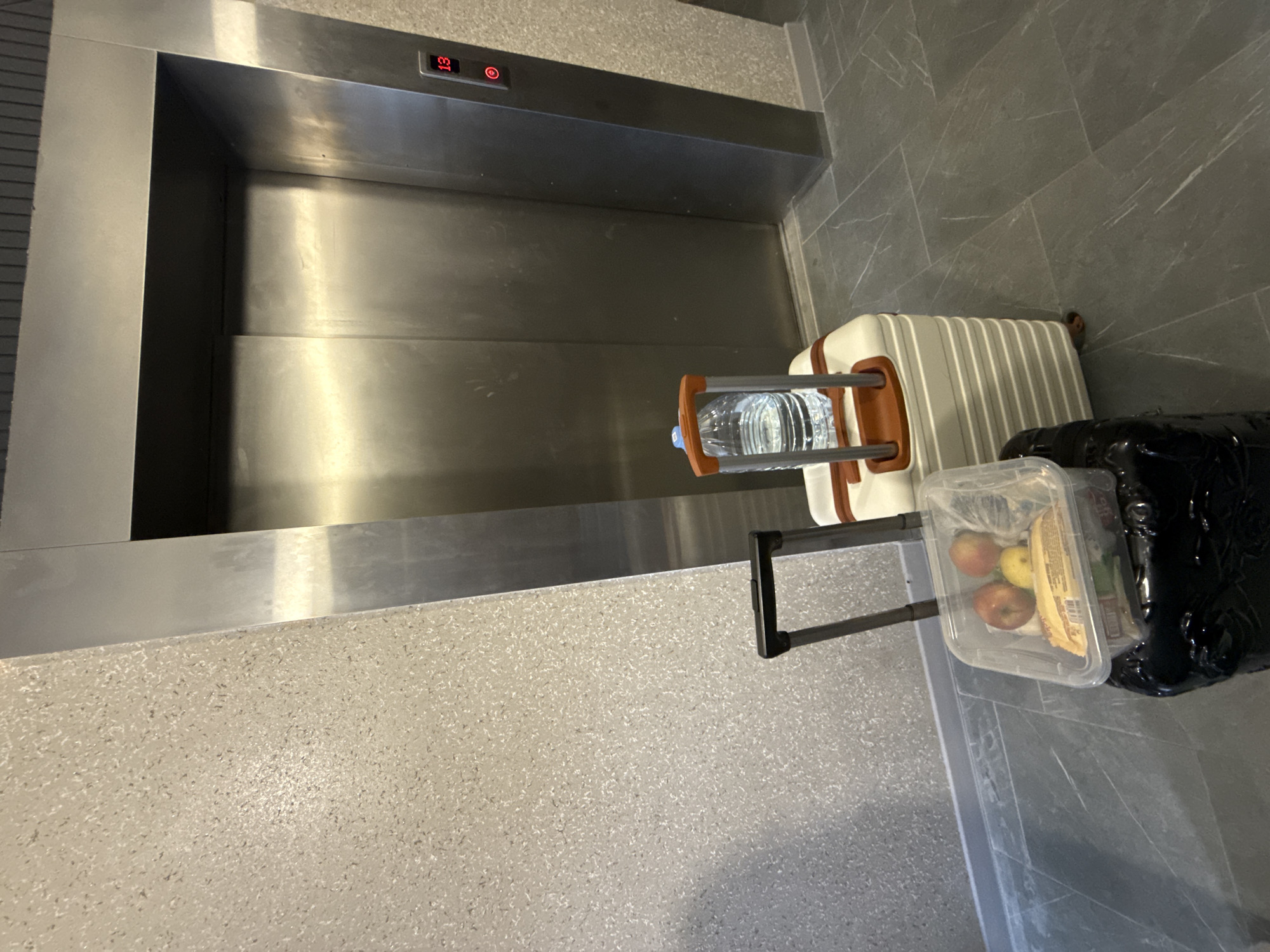 The image shows an elevator interior with a control panel indicating floor 13. Adjacent to the elevator entrance, a white and tan suitcase with a water bottle on the handle rests on the floor. Beside the suitcase is a transparent plastic container with apples and a granola bar, and a black plastic bag. The background environment features light gray flooring and elevator walls. The location in the city of Ovoshchnoy, Russia is unknown.