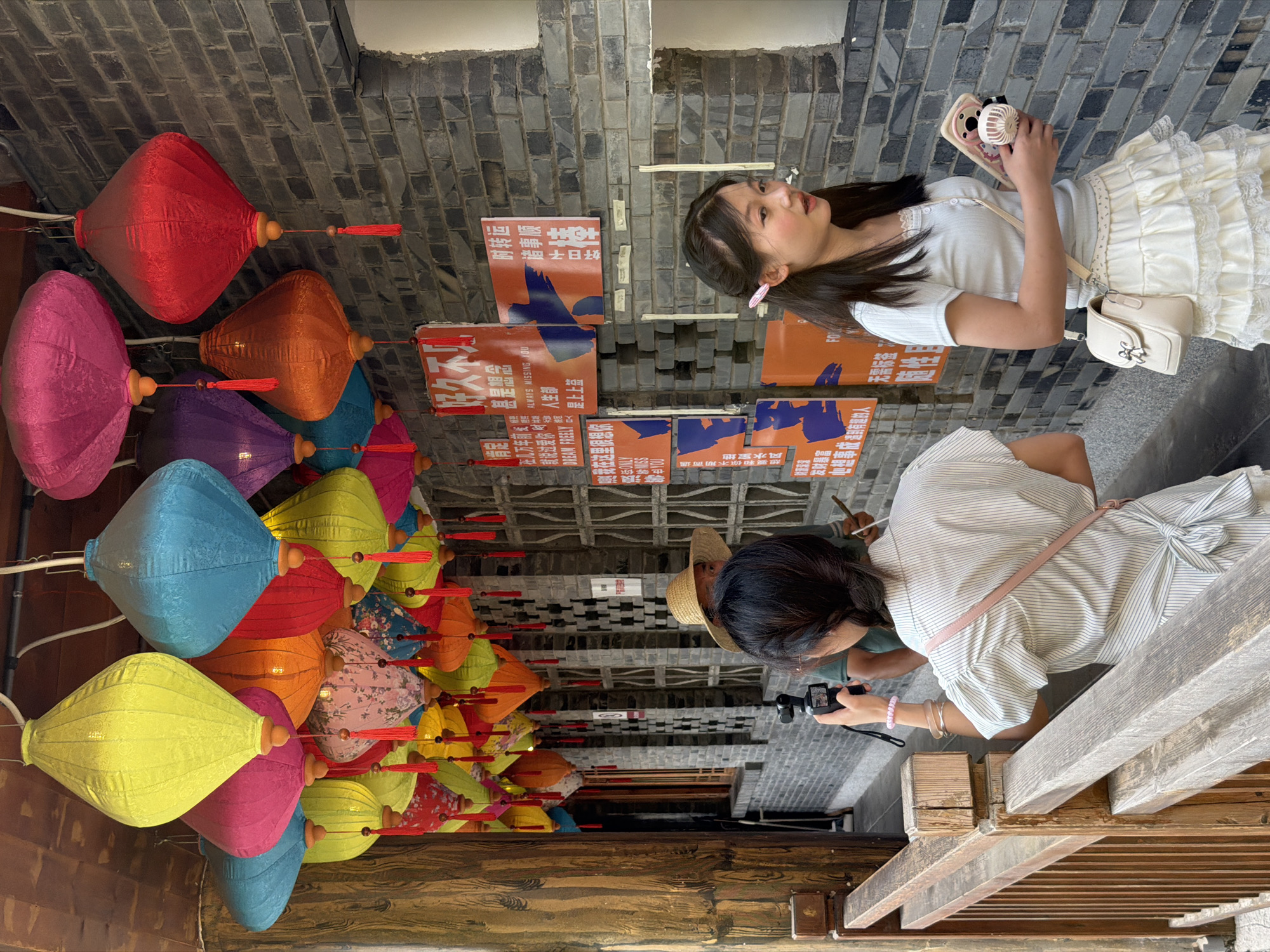 In Yuzhong District, China, a busy street scene unfolds under a canopy of colorful, hanging lanterns. Two young women are the central figures. The woman on the left, with her back to the camera, is capturing a video with a handheld camera, her hair in a braid. She wears a pinstripe shirt and a pink crossbody bag. To her right, the second woman, facing slightly away, has her hair in a partial ponytail with a pink clip. She wears a light blue short-sleeved top and a ruffled cream-colored skirt. She holds a smartphone with a fan attachment in her right hand and a light cream-colored shoulder bag. Behind them, a person wearing a straw hat is partially visible. The background features a brick wall adorned with orange and blue signage, some in Chinese characters. The environment suggests a vibrant, culturally rich area, possibly a market or tourist street, with traditional architectural elements and decorative elements.