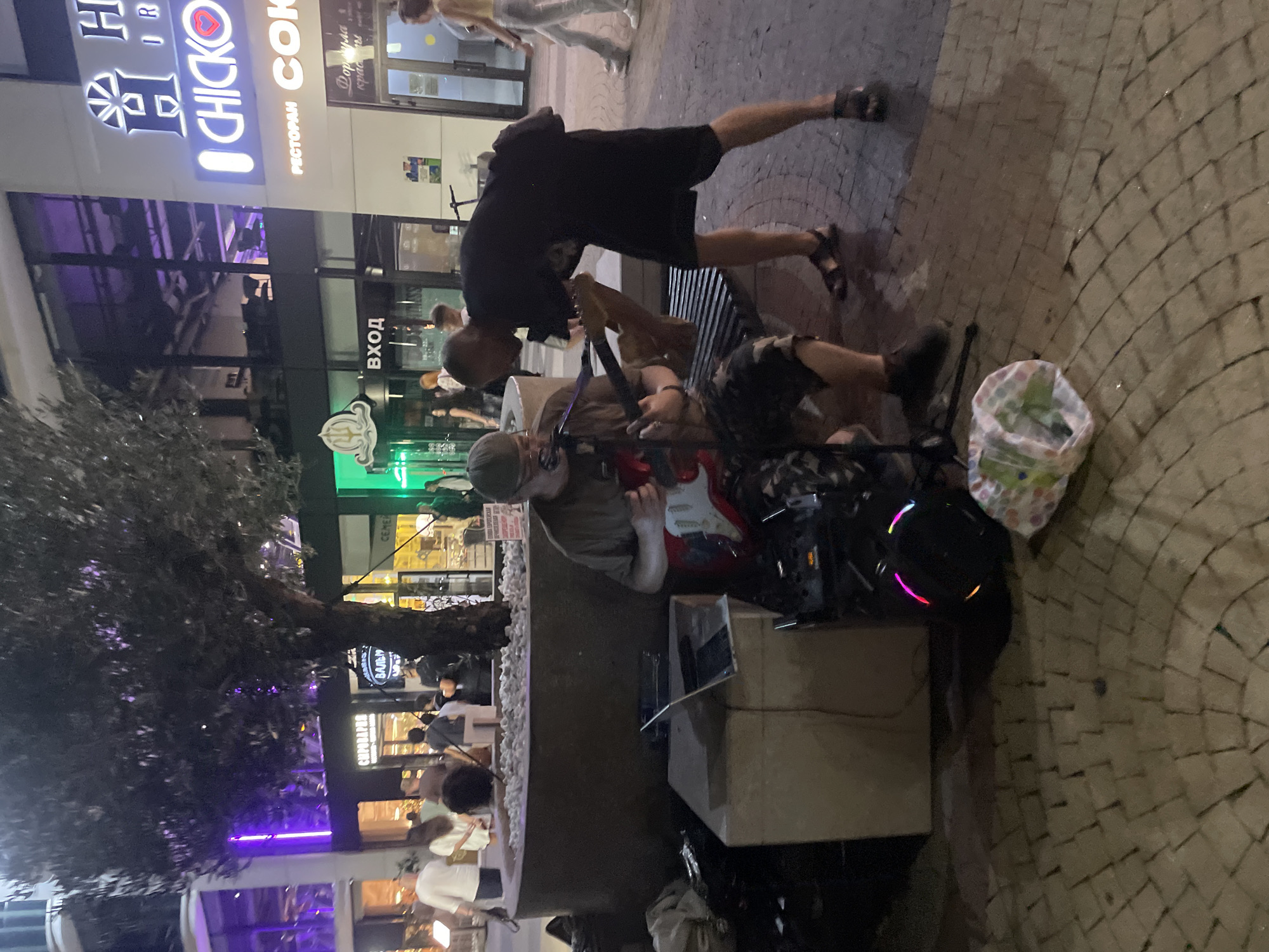 A street musician plays a red electric guitar on a paved area in front of a building complex at night. The musician is wearing a cap, glasses, and a t-shirt, and sits on a bench with camouflage-patterned pants. Another person stands behind him, also wearing dark clothing and sandals. In the background, there are shops and restaurants with illuminated signs. One sign reads "CHICKO" with a heart logo, and another says "РЕСТОРАН СОК". The ground is paved with cobblestones, and a tree with dark foliage is situated to the left of the frame. There is a laptop on a stone block next to the musician, and a speaker with colorful LED lights is on the ground. A plastic bag filled with items lies on the pavement near the speaker. People are walking in the background, some appearing to be customers or passersby. The scene suggests an urban environment at dusk or night, possibly a commercial or entertainment district. The location in Sadovyi, Russia, is inferred from the signage in Cyrillic, specifically "РЕСТОРАН СОК," which translates to "Restaurant Sok."
