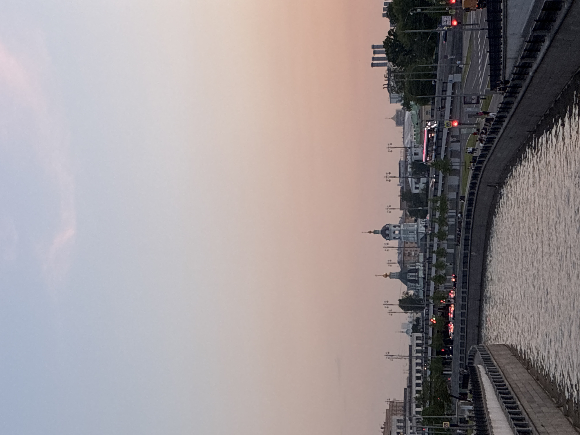 The image depicts a cityscape at dusk, likely along a river embankment in Перовское сельское поселение, Ukraine. A river, with a concrete embankment, stretches along the right side of the frame. On the opposite bank is a street with traffic lights showing red, indicating a busy urban area. Buildings of varying heights can be seen in the background, including what appears to be a church or cathedral with a dome. The sky transitions from a lighter color near the horizon to a deeper hue towards the top of the frame.