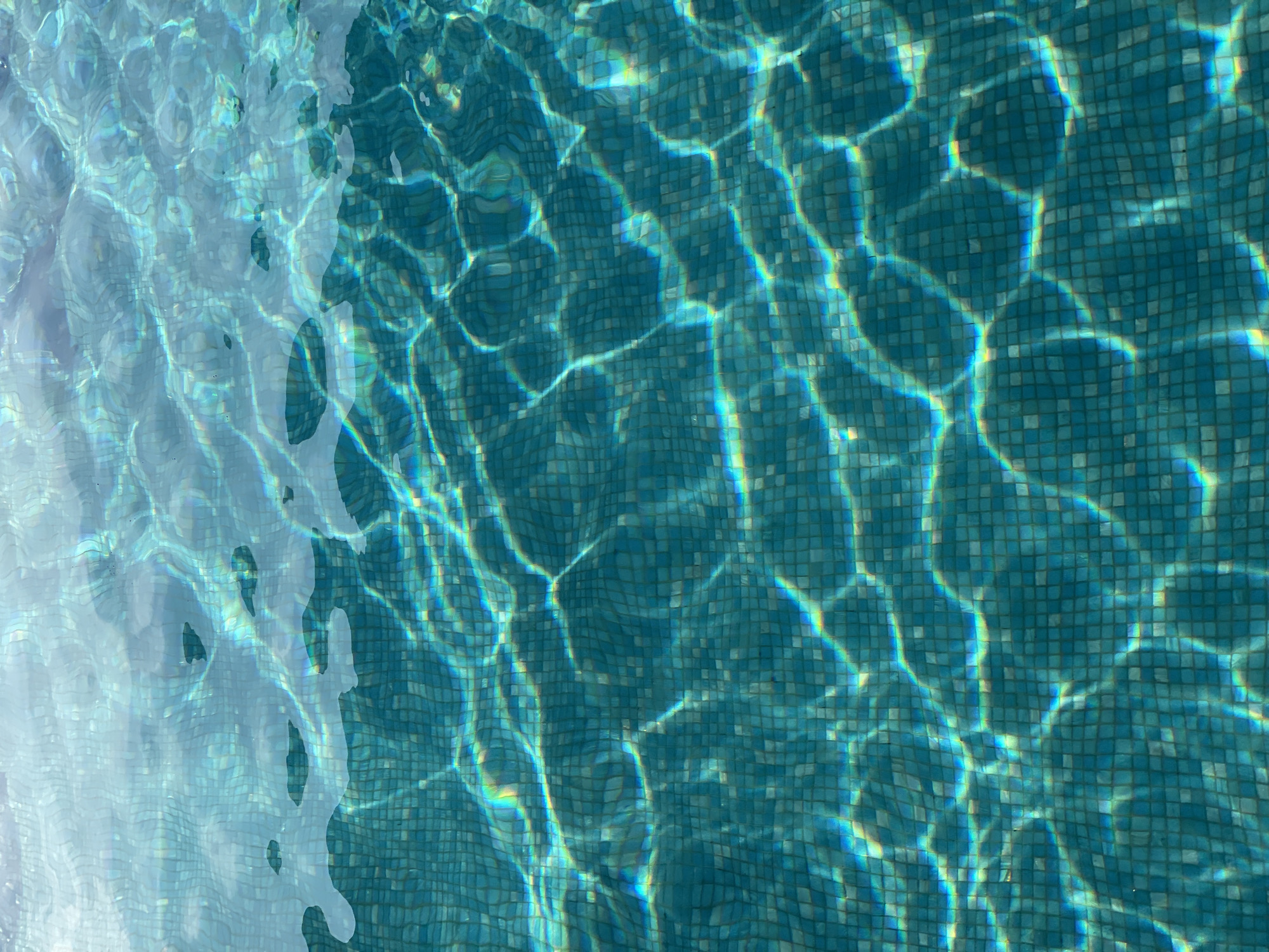 The image shows an overhead view of a swimming pool with turquoise tiles. Sunlight refracts through the water, creating shimmering patterns on the pool floor. One side of the frame shows clearer water reflecting more light, while the rest of the frame shows deeper water with stronger refraction patterns. There is no evidence to support that this photo was taken in the city of Central Jakarta, Indonesia.