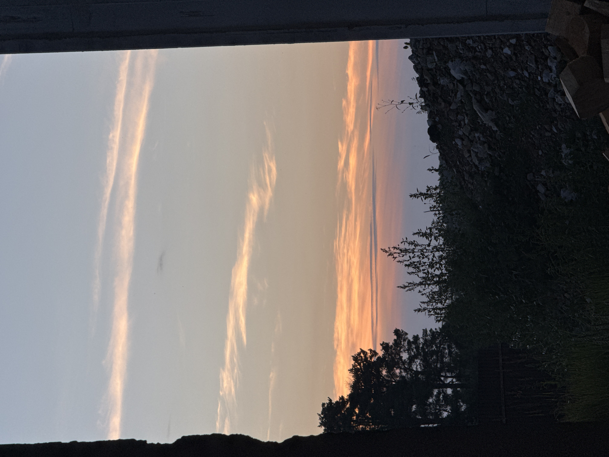 The image shows a twilight sky with wispy clouds tinged with pink and orange hues. In the foreground, silhouetted trees and dense foliage are visible. To the right, a concrete structure partially frames the scene, and a pile of rocks and debris is present. The location is within the Уздзенскі сельскі Савет, Belarus, suggesting a rural or semi-rural setting. No people or specific actions are depicted. The scene appears to be an outdoor environment during dusk.