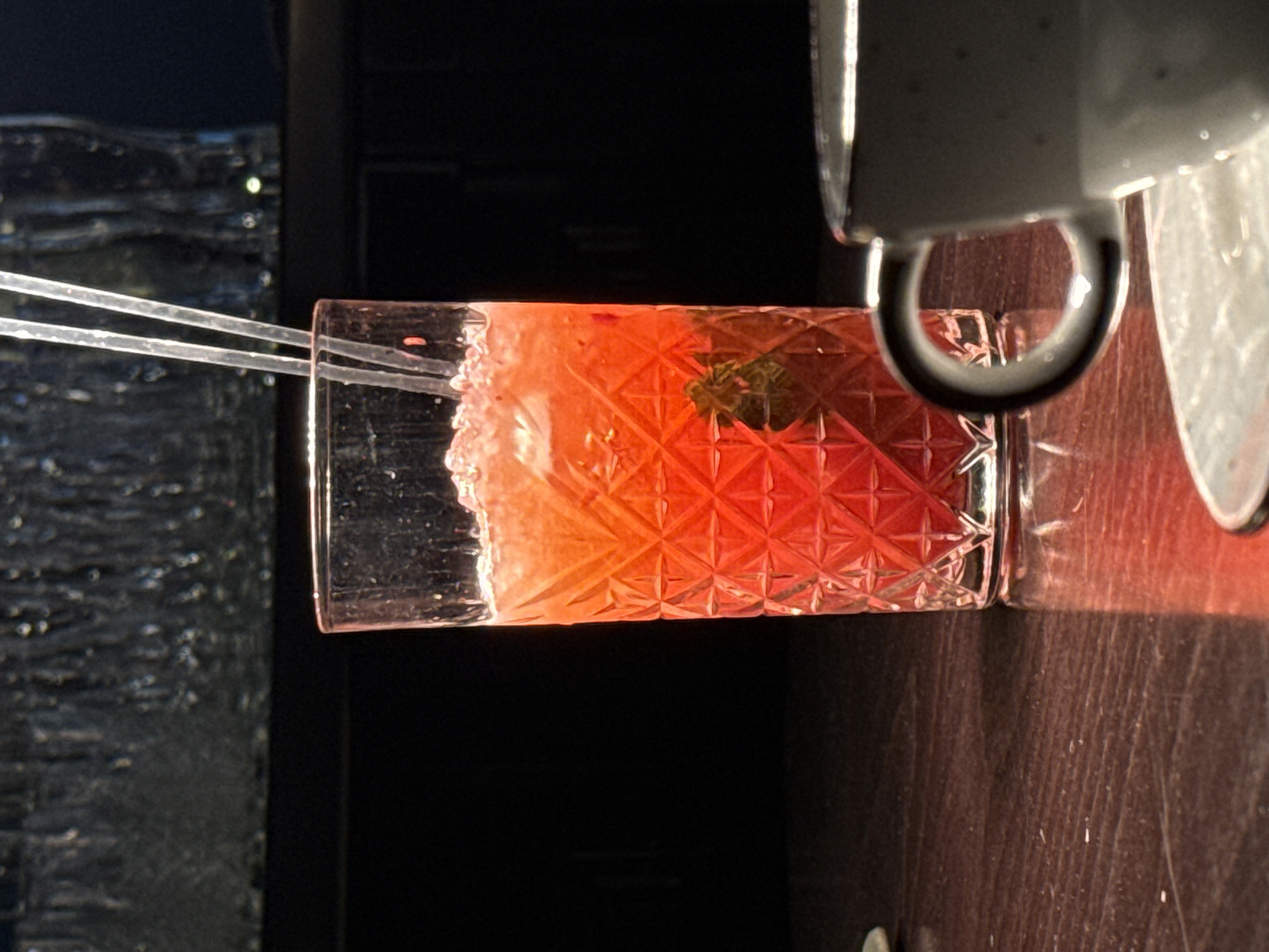 The image presents a brightly lit, close-up view of a cocktail in a patterned glass. The cocktail is a vibrant red color and appears to be garnished with a small green sprig. Two clear straws protrude from the drink. The glass sits on a wooden surface with a coffee cup in the upper right corner of the frame. The background is dark, with a textured panel visible behind the drink. There is no information in the image to identify its location in the city of Kursk, Russia.