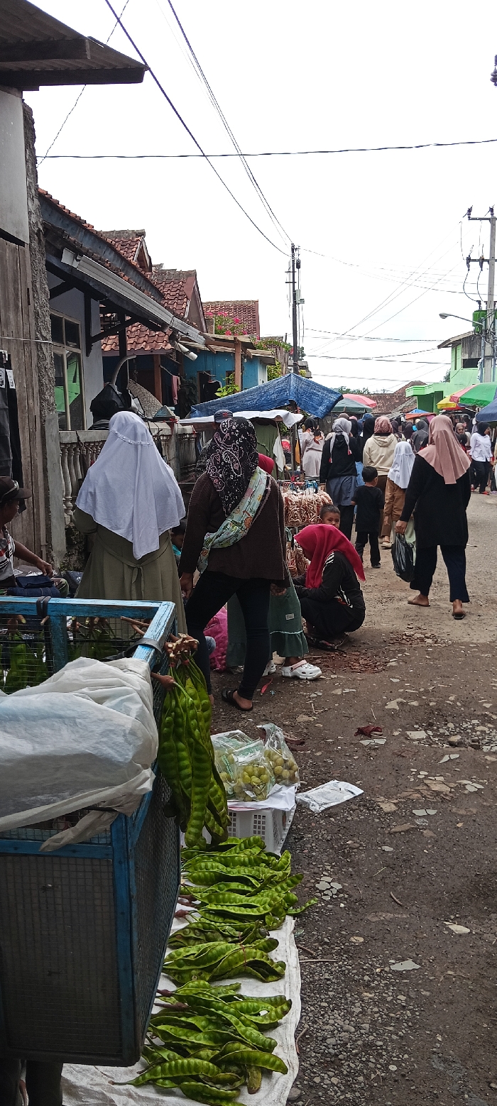 The scene takes place on a street in Kutawaringin, Indonesia, with the coordinates -6.974 latitude and 107.519 longitude. Several people are visible, some dressed in traditional clothing, such as headscarves. There are several objects in the scene, including a blue metal cage filled with green vegetable pods arranged on a white cloth, a bag filled with the same pods, a bag of small round fruit, and a few other bags. Several people are walking around, likely buying and selling these items, while others stand, wait, or sit. Several buildings line the street and there are overhead power lines. The street is paved with a mix of gray gravel and pavement.
