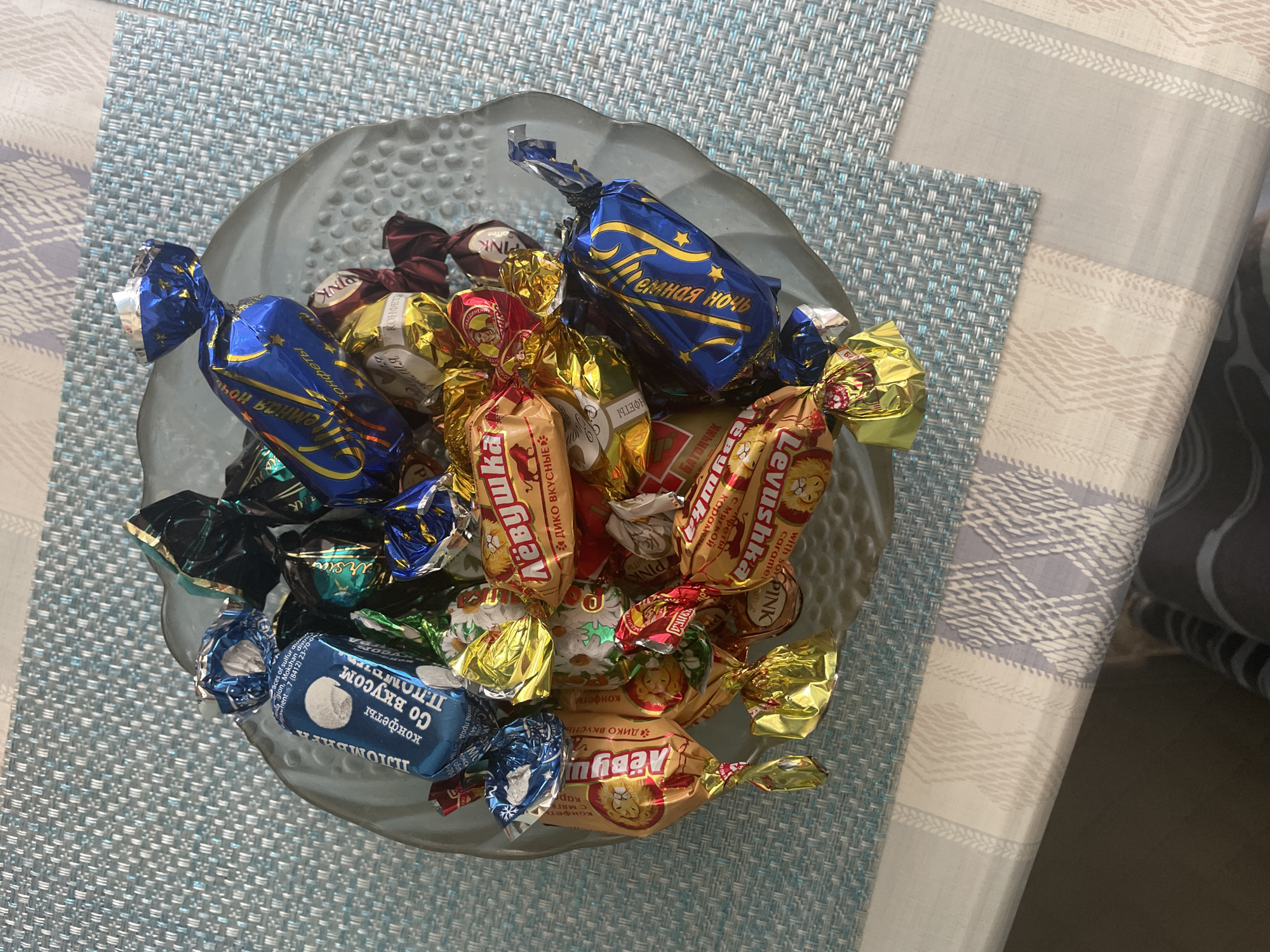 A close-up, overhead shot captures a glass bowl filled with an assortment of individually wrapped candies. The candies are presented on a table covered with a light blue woven placemat that has a subtle striped pattern. The bowl itself is made of clear, textured glass with an organic, slightly rippled edge. The candies, with their crinkled foil wrappers in various colors including blue, gold, red, and green, are densely packed within the bowl. Some of the wrappers display text in what appears to be Cyrillic script, suggesting Russian origins. The ambient lighting casts soft shadows, highlighting the reflective surfaces of the wrappers and the texture of the placemat. There are no people visible in the frame, and the scene is static, focusing solely on the arrangement of the candies. The image does not provide any specific visual cues to pinpoint its exact location in the city of Chaikovsky, Russia, beyond the general context of a domestic or retail setting where such candies might be found.
