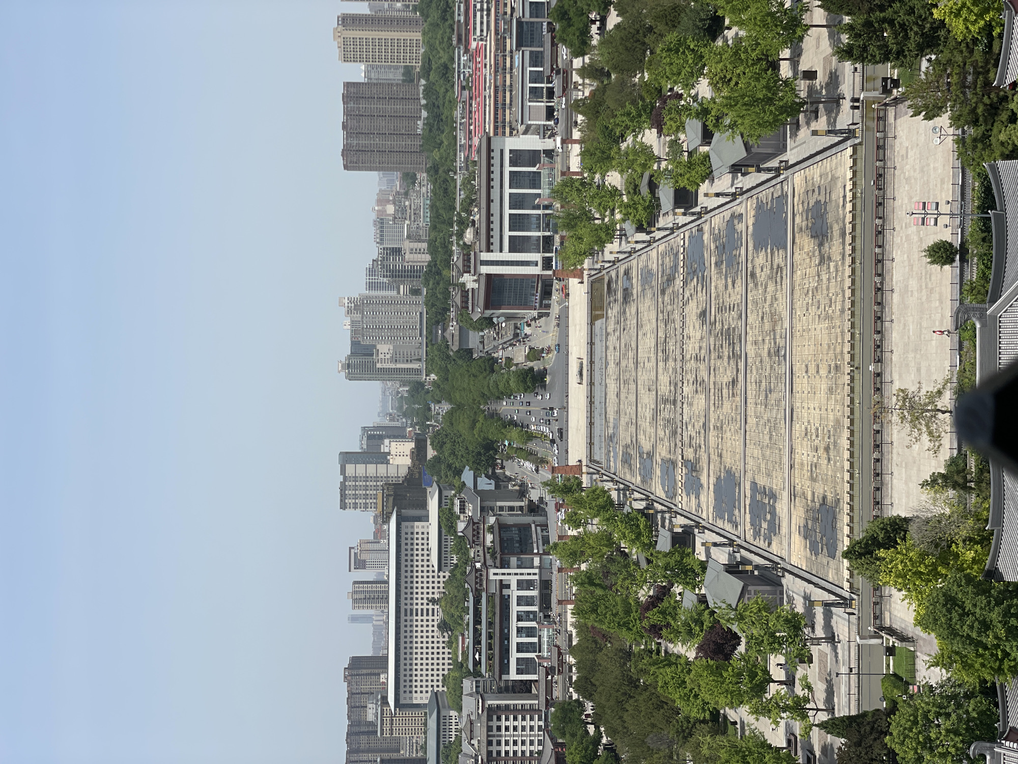 An aerial view depicts a city skyline with a combination of high-rise buildings and traditional Chinese architecture in Yanta District, China. A large, rectangular structure with a patterned surface dominates the foreground, potentially part of a historical site. The street below is lined with trees. Numerous buildings of varying heights and designs are in the background under a muted sky. The scene is set during daylight hours.