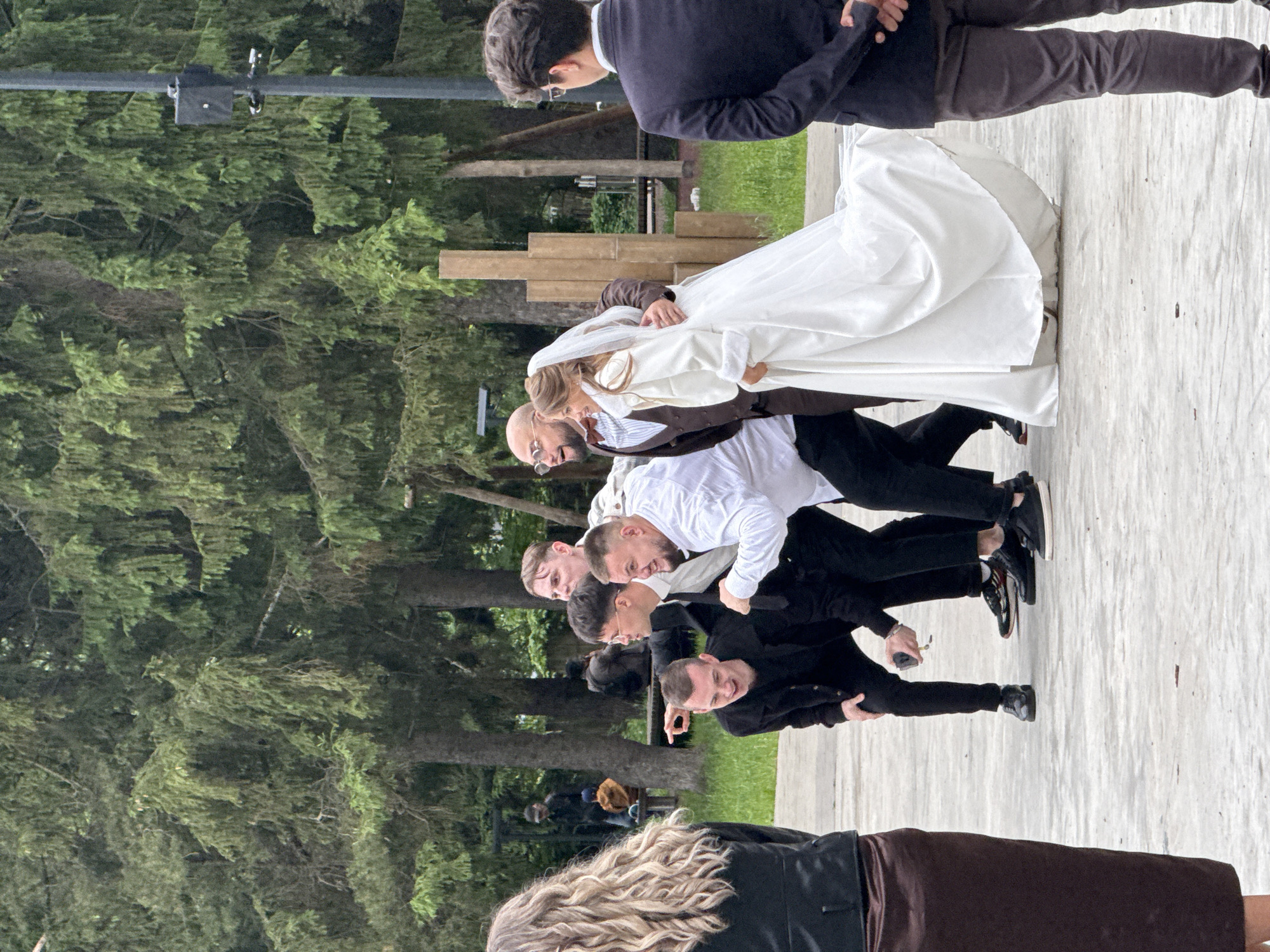 The image depicts a daytime outdoor scene in Votkinsk, Russia, featuring a group of eight individuals on a paved surface against a backdrop of dense evergreen trees. The lighting suggests an overcast sky.
In the foreground, a woman stands with her back to the viewer on the far left. She has long, wavy blonde hair and wears a black jacket over a dark brown skirt. On the far right, a man also stands with his back to the viewer, wearing a dark suit jacket, light shirt, and dark trousers, with his hands clasped behind his back. He wears glasses.
The central group consists of six individuals, appearing to interact or pose. From left to right within this group:
1. A young male with short dark hair, wearing a black jacket and black pants, is squatting. He holds a light-colored object, possibly a smartphone, in his right hand and points with his left hand towards the left. His mouth is slightly open.
2. A young male with short dark hair and glasses, wearing a black suit jacket, white shirt, and black tie, leans forward, pointing with his right index finger towards the left and looking in that direction with an open mouth.
3. Partially visible behind the previous two, a young male with short light-brown hair wears a white shirt.
4. A male with short dark hair, wearing a white long-ssleeved shirt and dark pants, has his left arm bent with a clenched fist, looking animated towards the left.
5. A bald male with a beard and round glasses, wearing a brown suit and red bow tie, has his left arm around the waist of the woman beside him. He looks towards the left with a slight smile.
6. A female, presumably a bride, wears a long white wedding gown with a white long-sleeved coat over it and a white veil. She holds her dress with her right hand and smiles while looking towards the left.
The ground is a light-colored paved surface. In the background, numerous tall, dark green coniferous trees are visible. Behind the central group, several vertical wooden posts or sculptures are arranged. On the right side of the image, a dark utility pole stands, equipped with a black box and two security cameras. In the distant background, through the trees, faint glimpses of other structures, including what appears to be a park bench, are present.