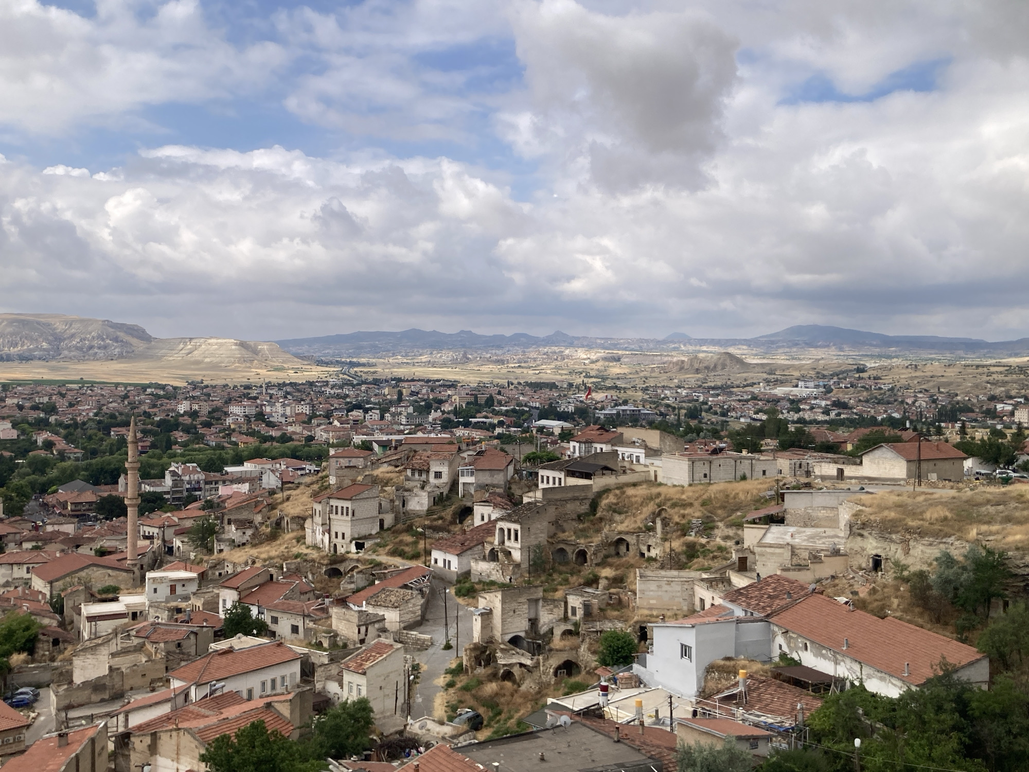 This is an eye-level, medium-shot, outdoor, long-distance photograph of the city of Avanos İlçe Merkezi, Turkey. The image is captured on a partly cloudy day, with white and gray clouds dominating the upper portion of the frame, interspersed with patches of blue sky. In the middle ground, the city sprawls across rolling hills, characterized by a dense collection of buildings with terracotta roofs. Many structures appear to be made of stone or concrete, with some exhibiting a rustic, traditional architectural style, including cave dwellings carved into the hillsides.
The foreground and mid-ground are dominated by this dense urban landscape. Prominent features include a tall, slender minaret from a mosque, several multi-story buildings, and numerous smaller houses. The terrain is dry and somewhat arid, with patches of dry grass and scrub vegetation visible between the buildings and on the hillsides. Roads wind through the city, and a few vehicles are discernible.
In the far distance, a range of hills and mountains form the horizon, appearing hazy and muted under the atmospheric conditions. The overall impression is of a historic and densely populated settlement nestled within a unique geological landscape. No people are clearly visible in the image. The light is diffused, suggesting it is not direct midday sun.